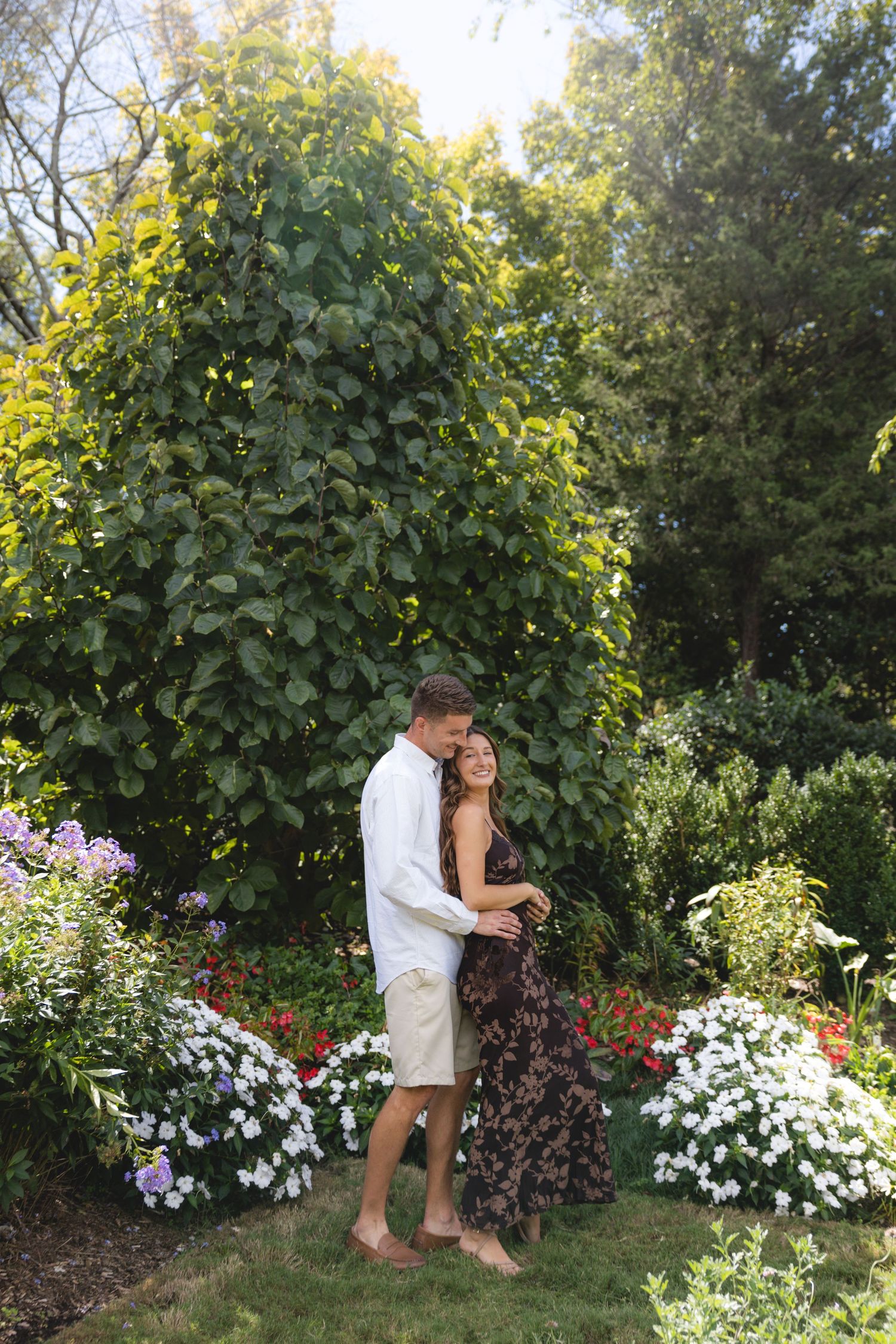 Couple embracing in a lush garden surrounded by colorful flowers and green trees on a sunny day.