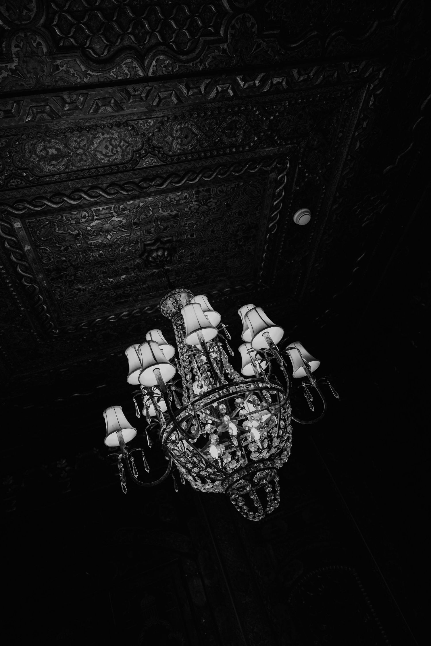 Black and white photo of an ornate crystal chandelier with lampshades hanging from a decorative carved ceiling.