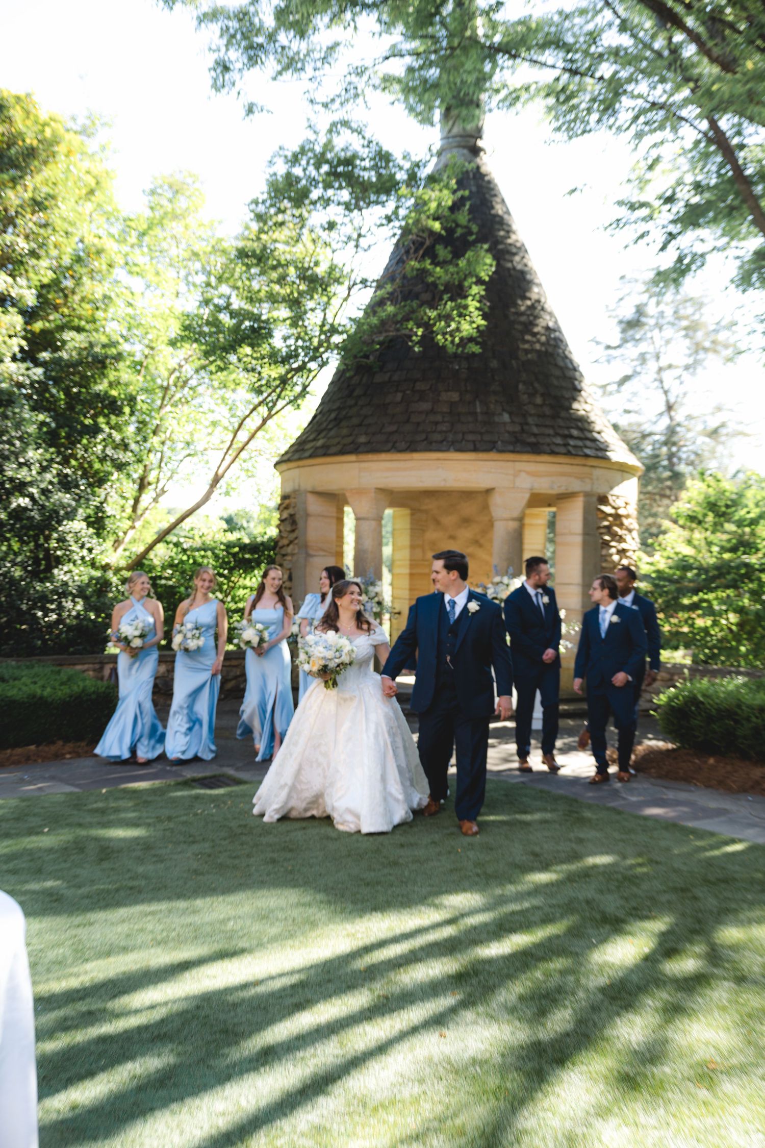 Bride and groom walk with wedding party in light blue dresses and navy suits near a stone gazebo in a lush garden.
