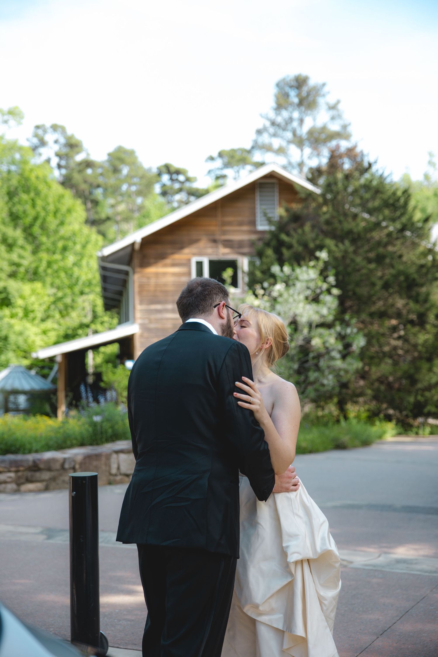 Bride and groom sharing a kiss outdoors near a rustic wooden building surrounded by lush green trees on their wedding day.