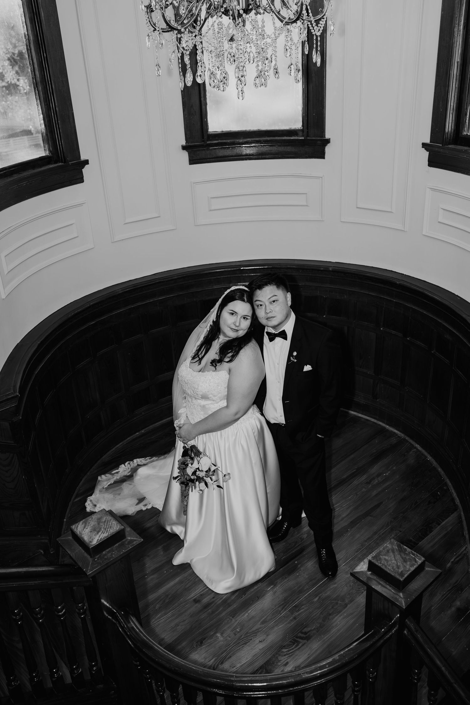 Black and white wedding portrait of a bride in a veil and a groom in a bow tie embracing on a curved staircase landing.