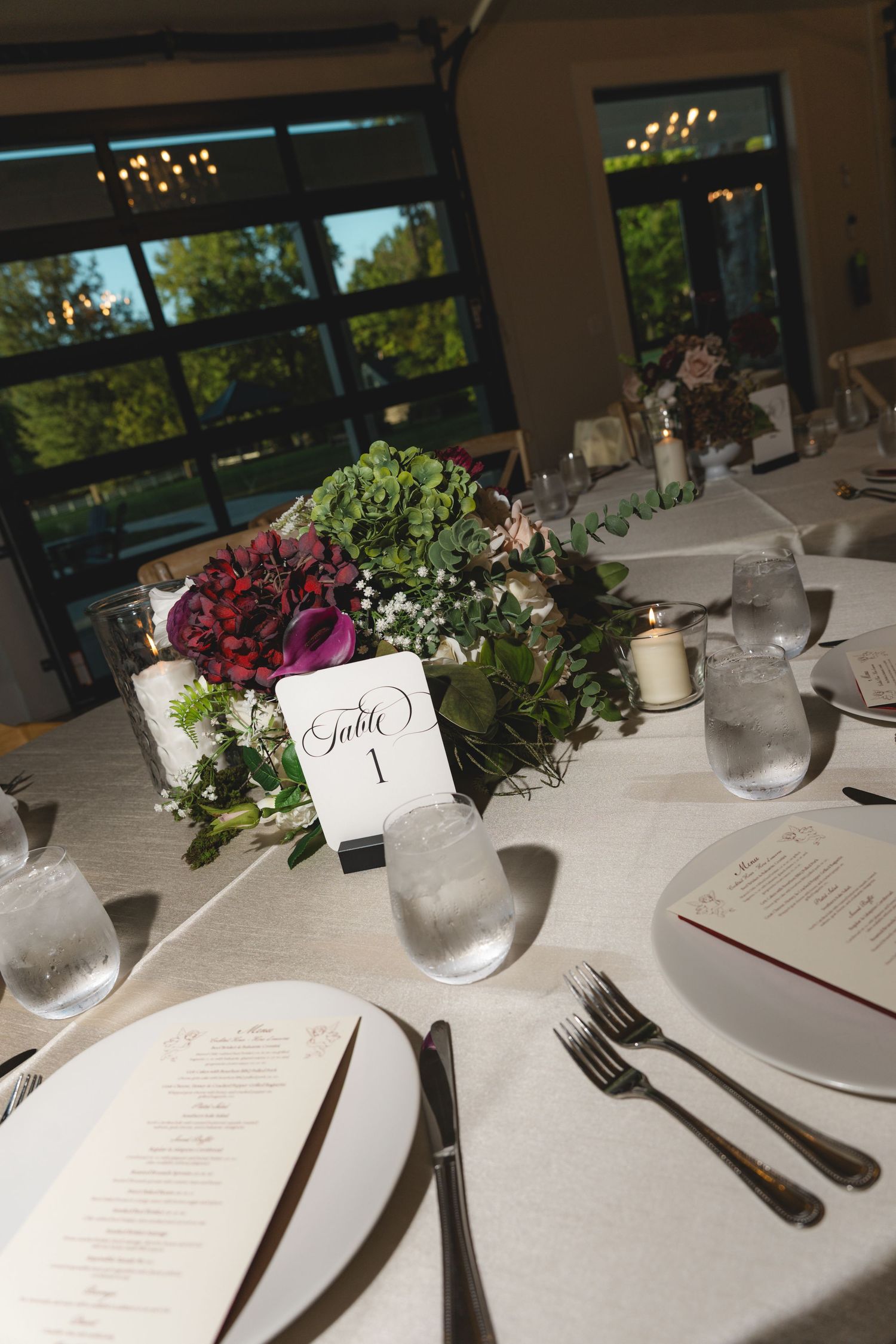 Wedding reception table 1 with a lush floral centerpiece, candles, menus on white plates, and glassware beside large windows.