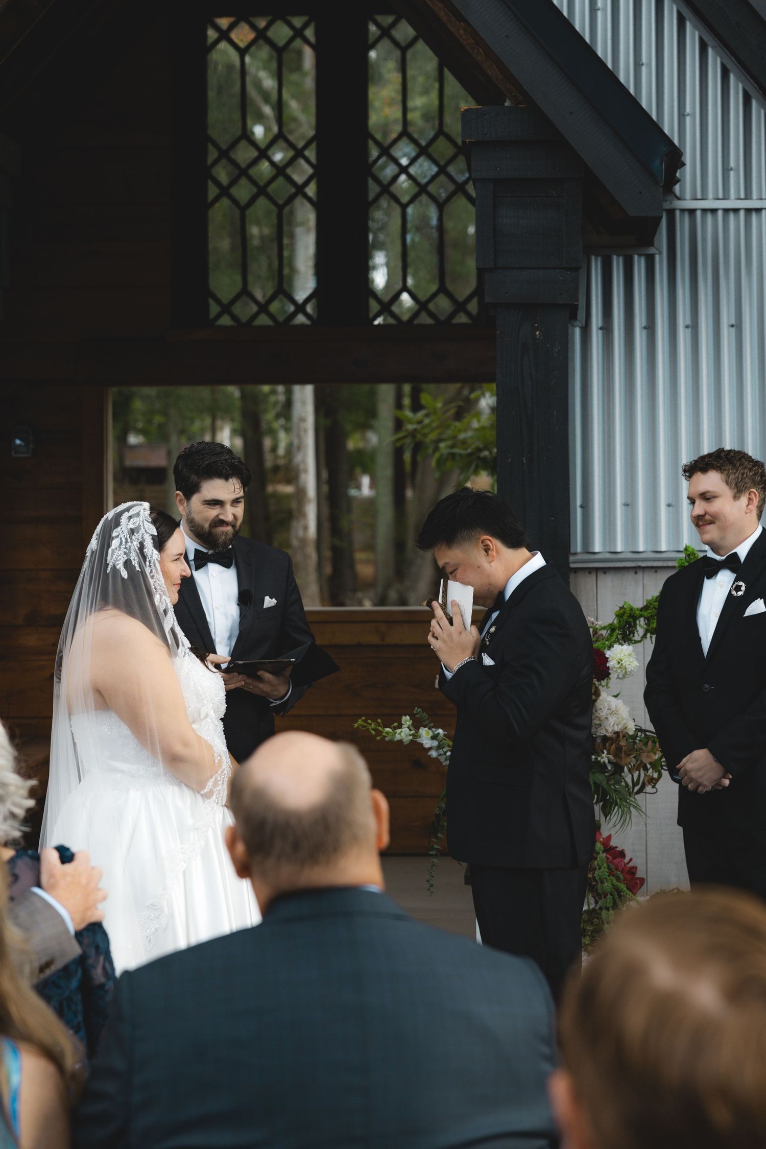 Bride in a lace veil and groom wiping tears at an outdoor wedding ceremony as the officiant reads and guests watch.
