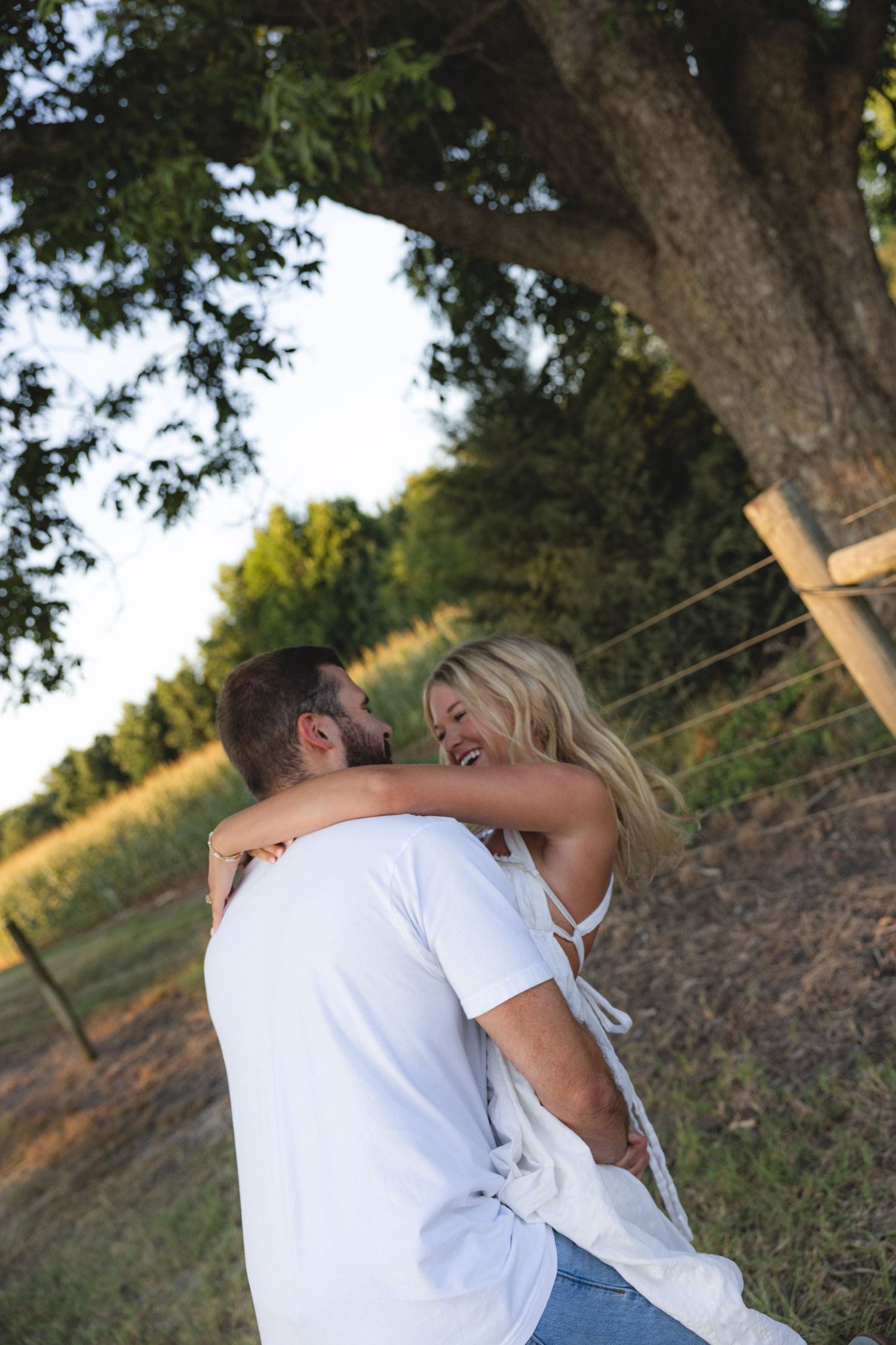 A couple embracing and laughing together outdoors near a large tree in a sunny rural field.