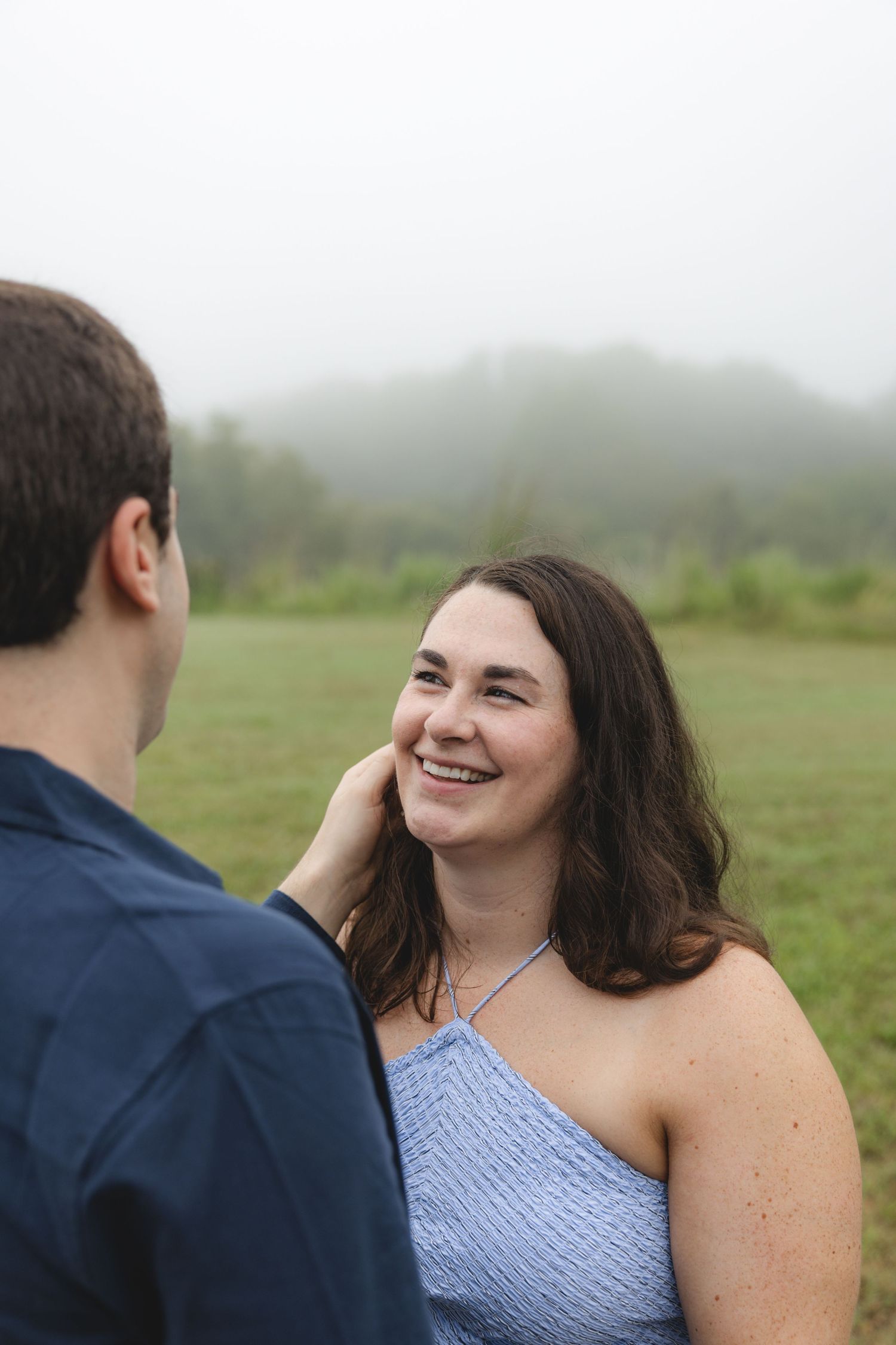 A smiling woman in a blue top gazes up at a man touching her face, with a misty green field in the background.