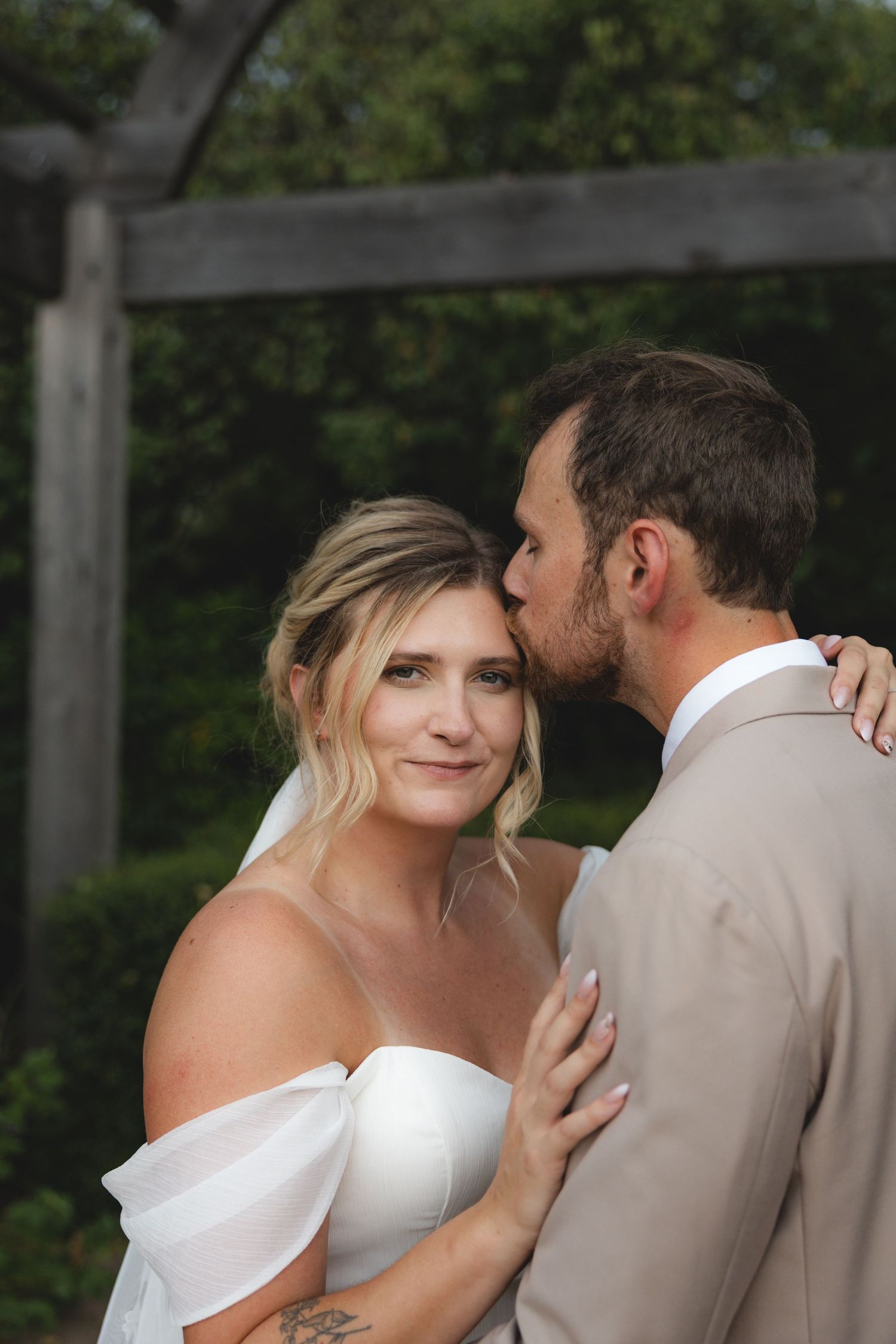 Groom in tan suit kissing bride in off-shoulder white gown outdoors near wooden arbor.