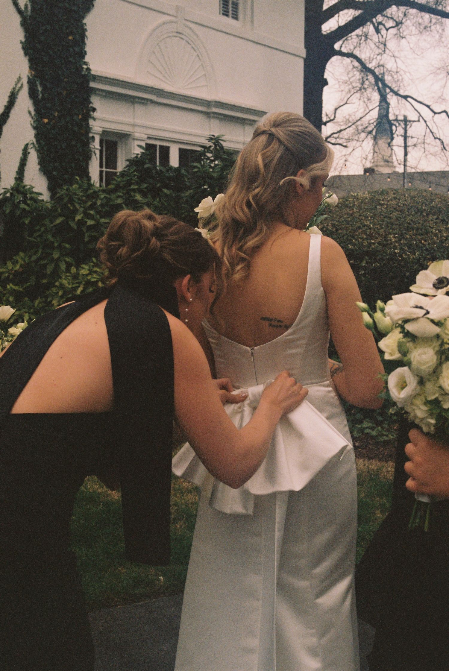 Two women in white dresses embrace in a garden with a building and trees in the background.