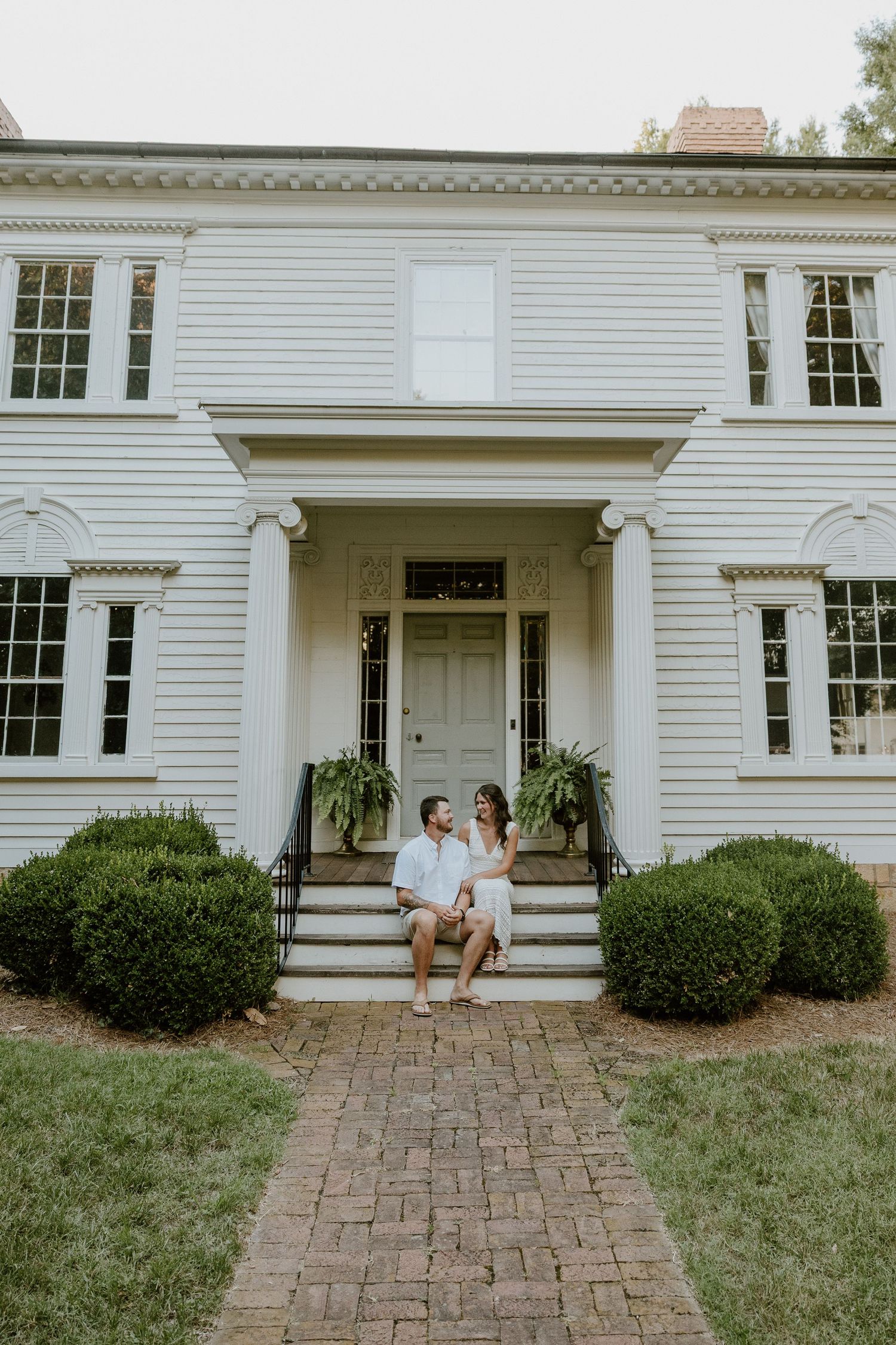A couple sits on the front steps of a classic white colonial style house.