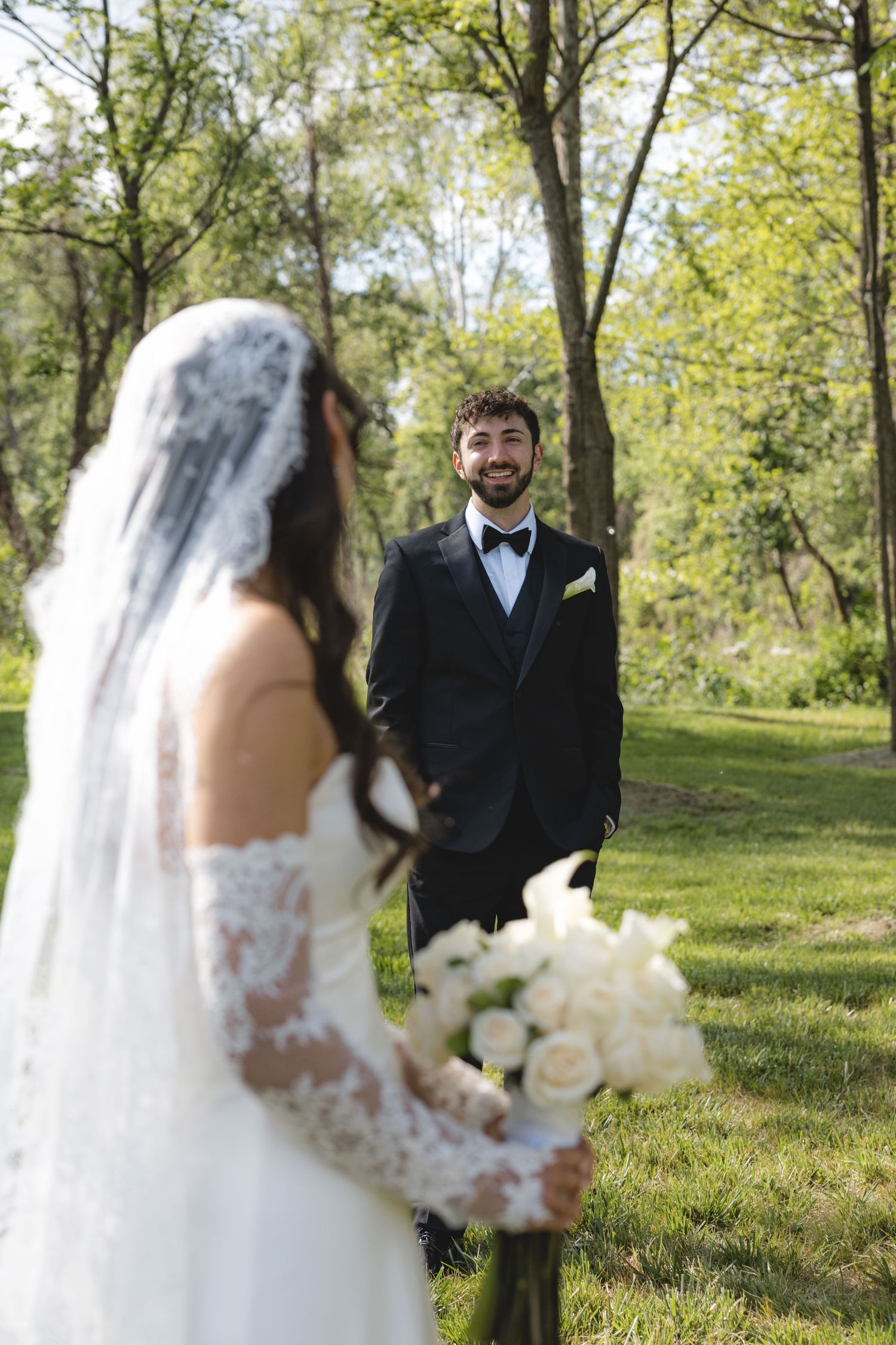 Smiling groom in black tuxedo reacts during first look with bride holding white rose bouquet.