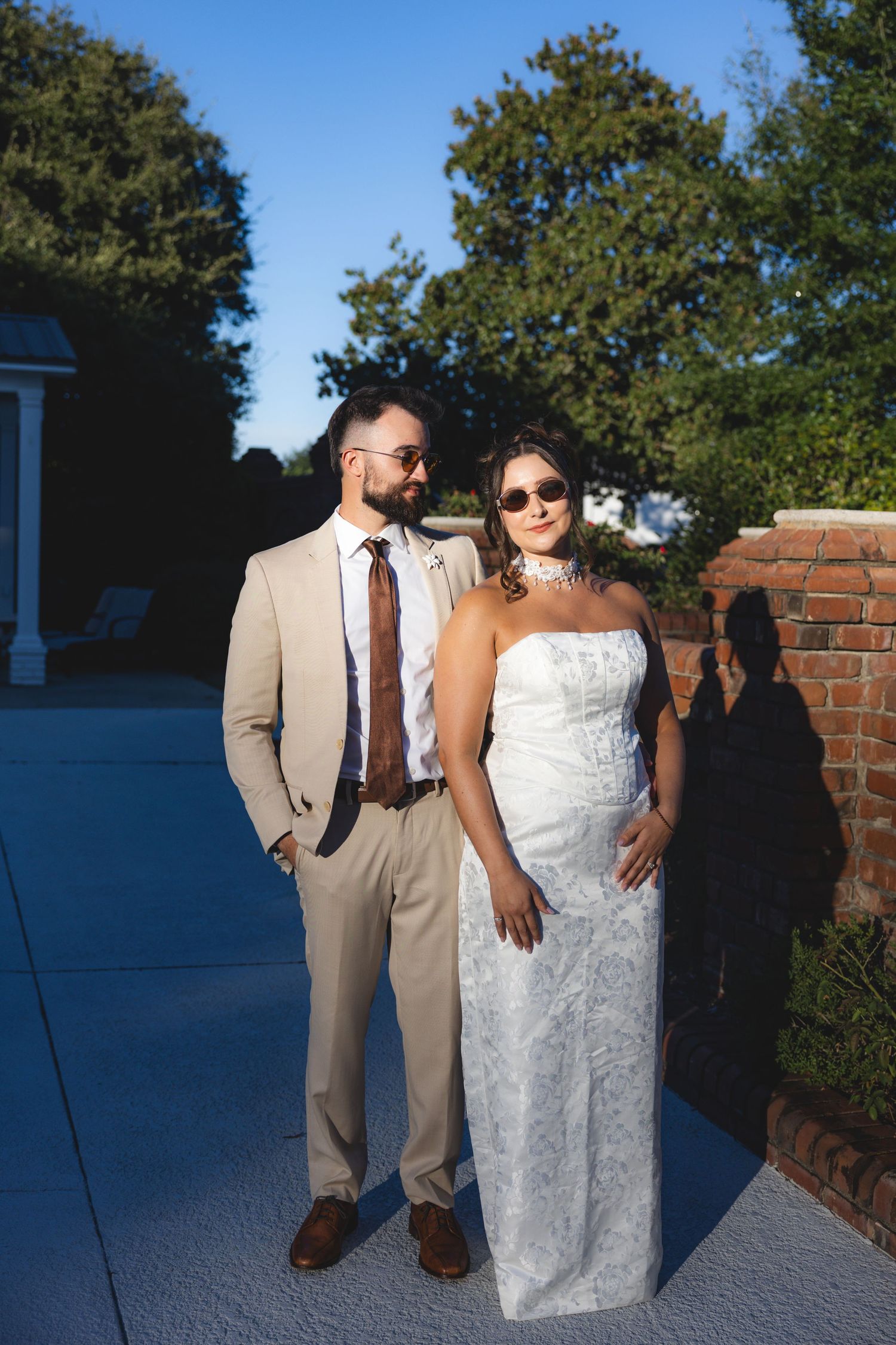 Bride in white strapless gown and groom in tan suit with brown tie pose outdoors on their wedding day.