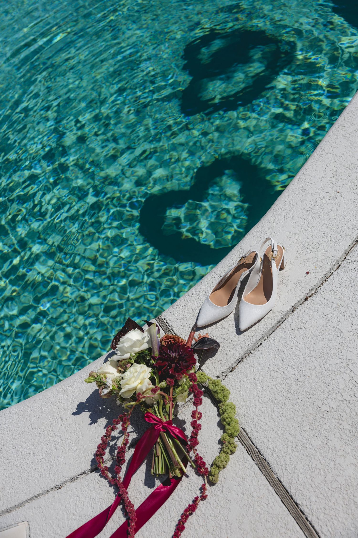Bridal bouquet and white heels by a pool with heart-shaped shadows on turquoise water.