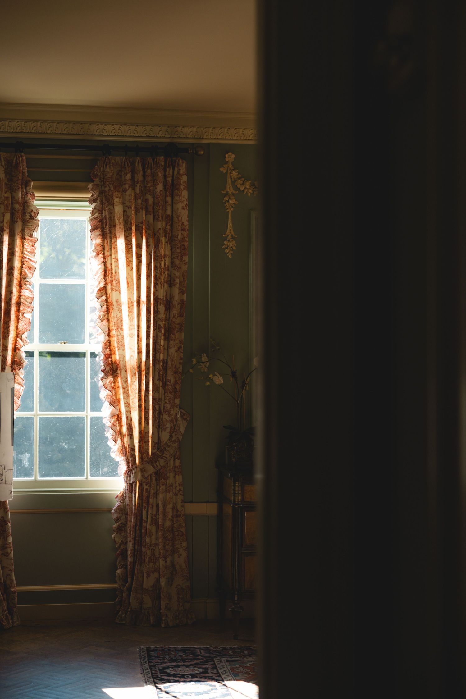 Elegant room with floral curtains framing a sunlit window, casting warm light on hardwood floors in a classic interior.