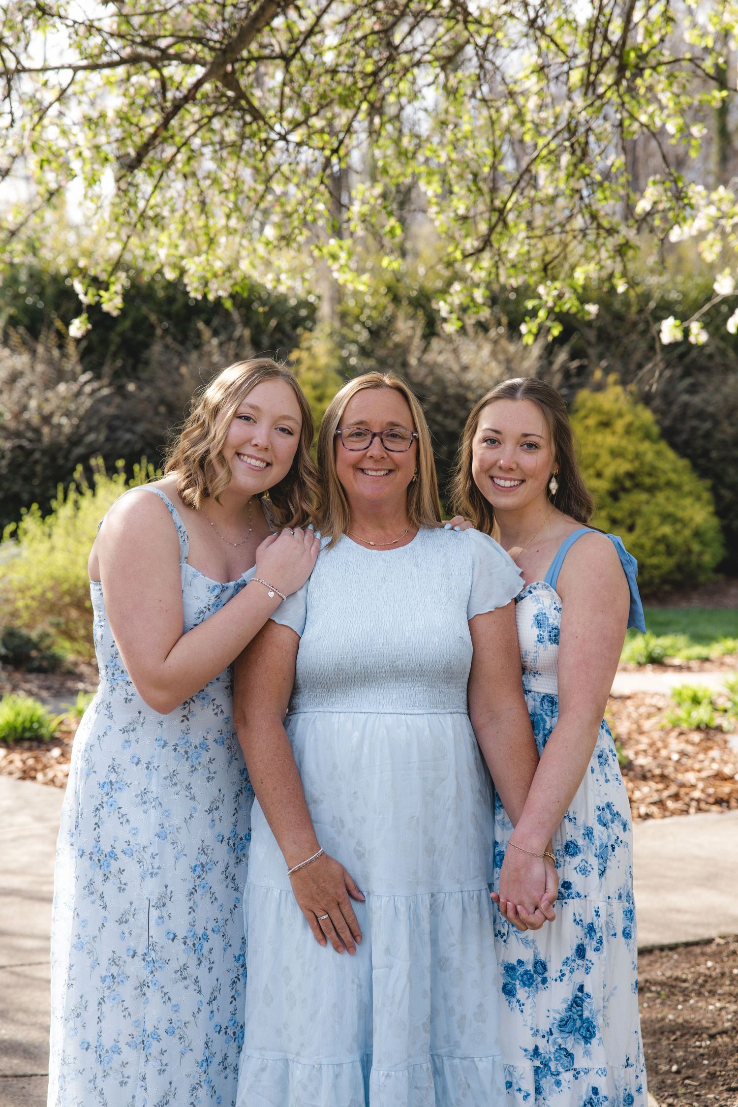 Three women in matching blue floral dresses smile together outdoors in a garden with blooming spring trees.