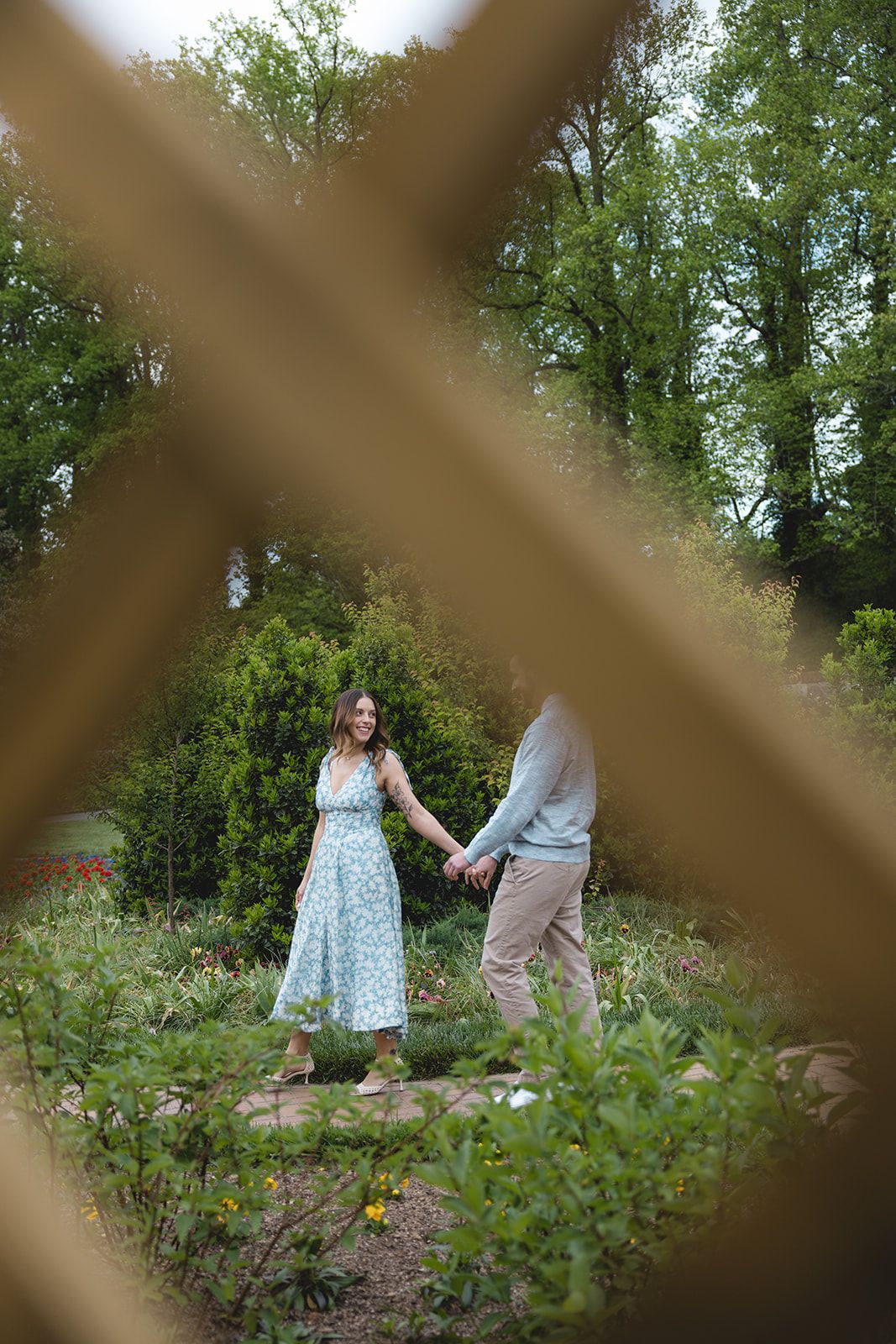 Couple holding hands in a lush garden, viewed through a wooden lattice fence, woman in floral dress.