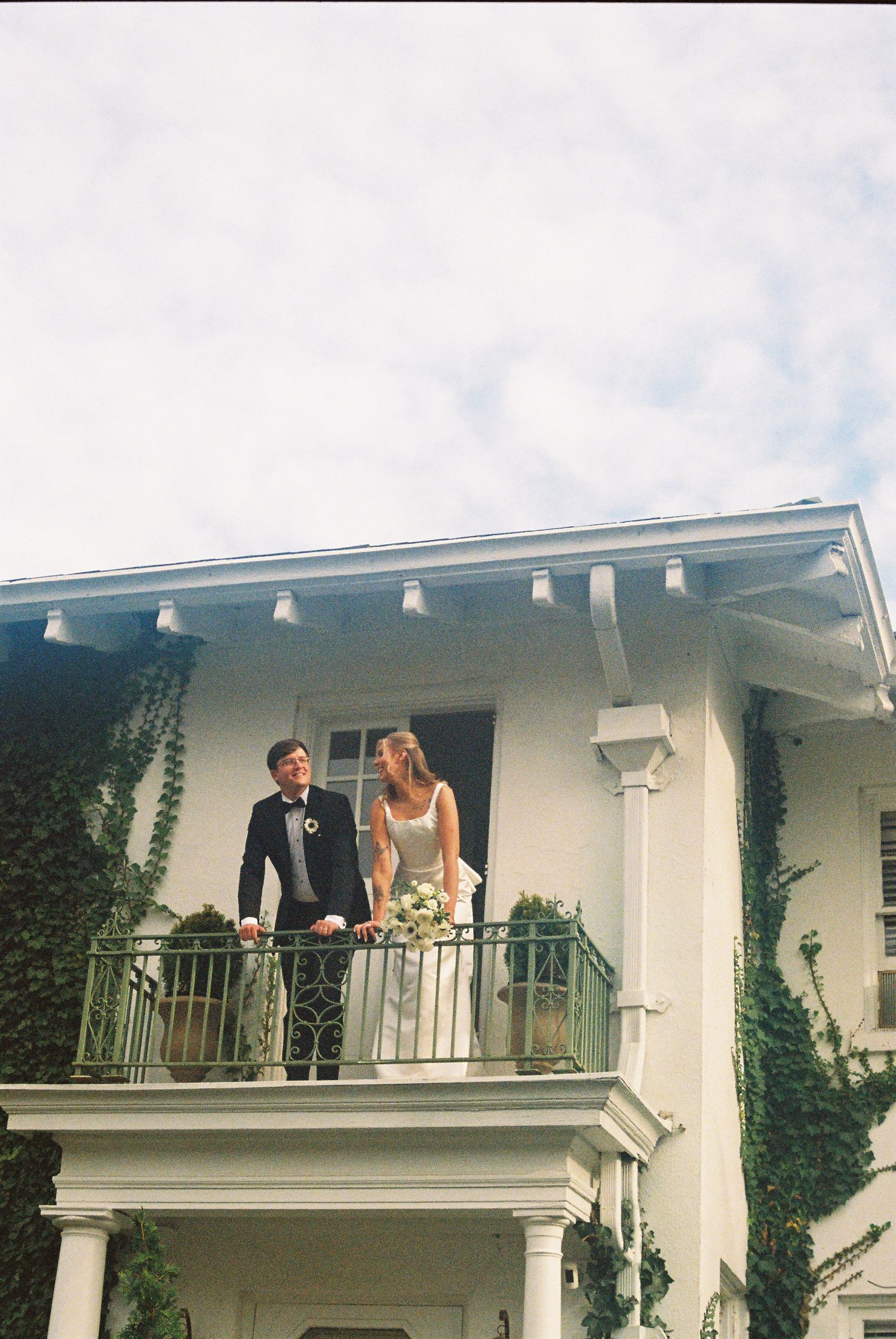 A bride and groom stand together on an ivy-covered white mansion balcony on their wedding day.
