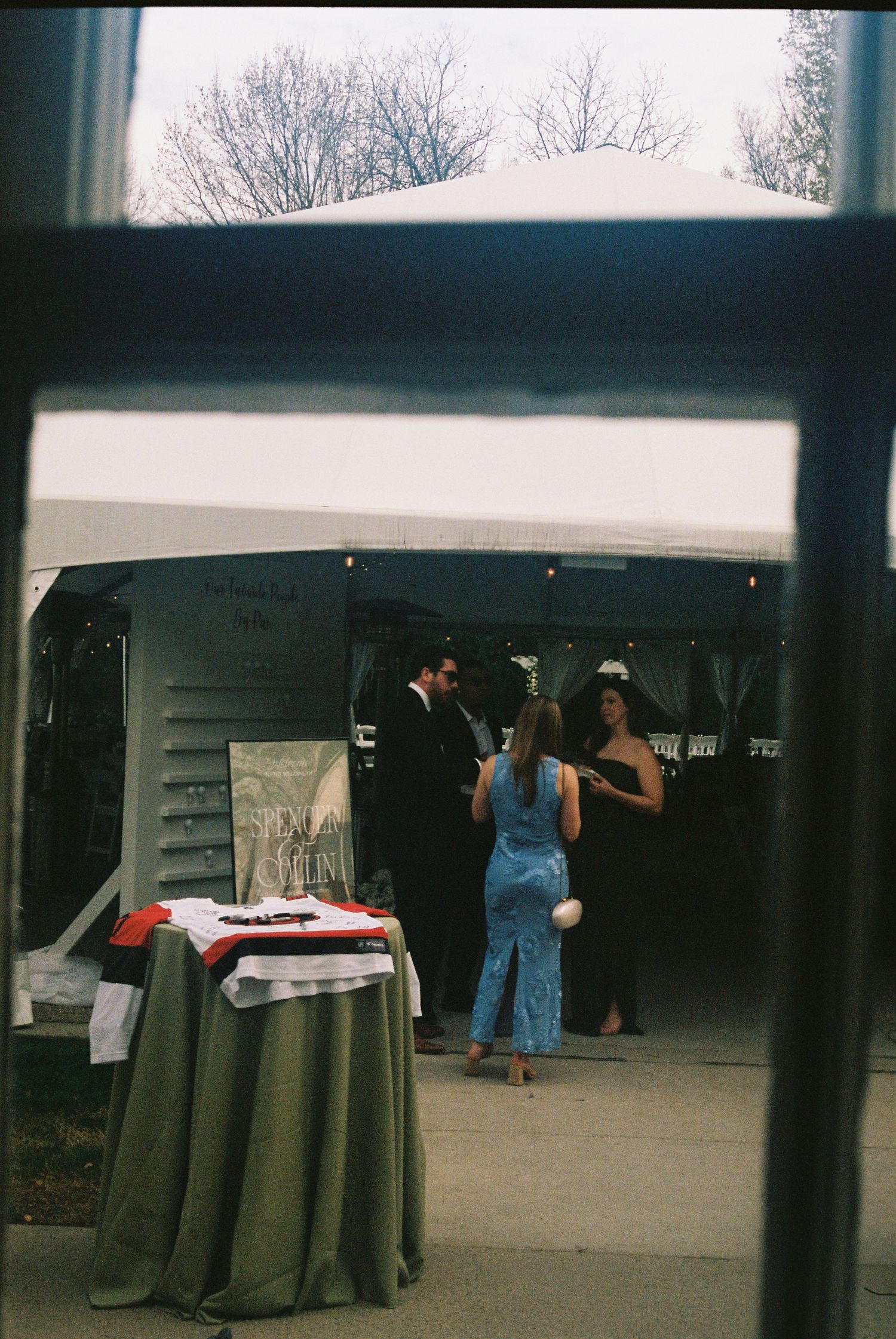 Guests in formal attire gather under a white tent at an outdoor wedding reception with draped tables nearby.