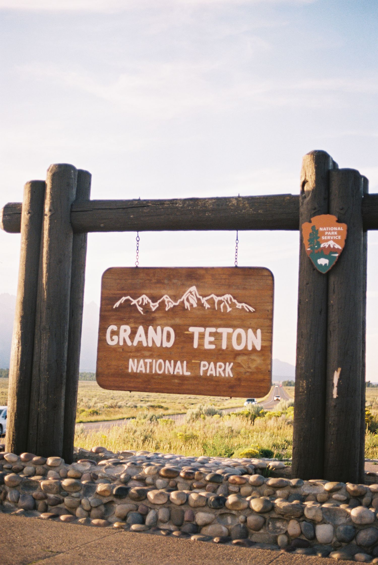 Wooden entrance sign for Grand Teton National Park with mountain engravings, set on a stone base under a partly cloudy sky.