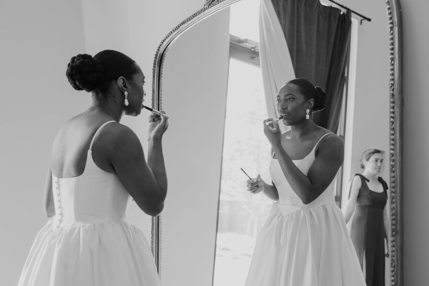 Black and white photo of a bride applying makeup in a mirror, with a bridesmaid visible in the background.