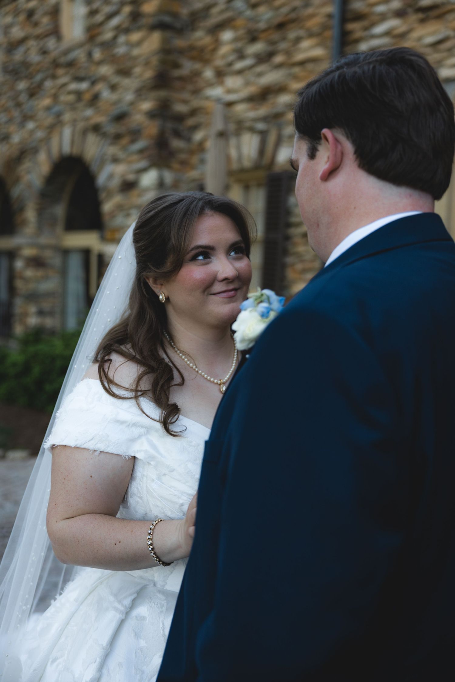 Bride in white gown and veil smiles lovingly at groom in navy suit outside stone building.