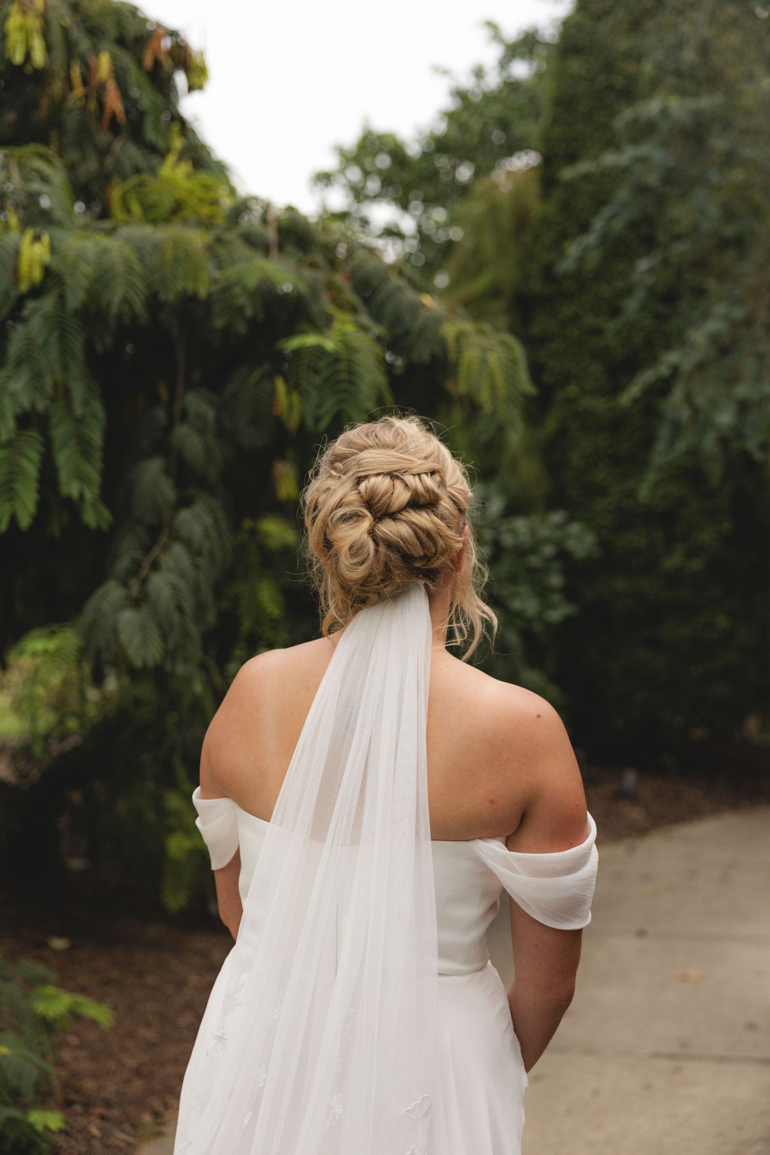 Bride seen from behind wearing off-shoulder white gown with elegant braided updo and flowing veil, surrounded by lush greenery.
