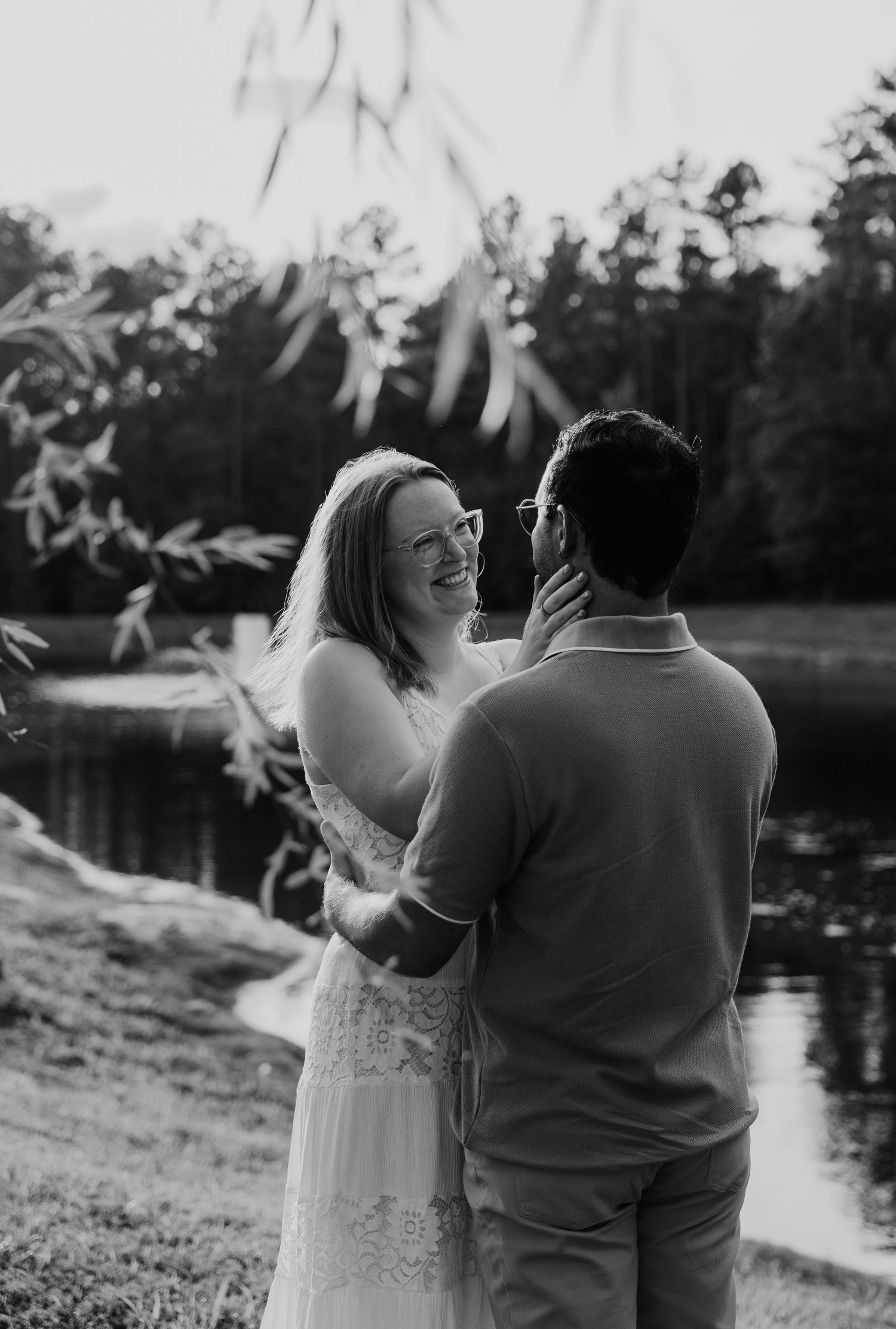 Black and white photo of a smiling bride in white dress gazing at groom near a pond surrounded by trees.