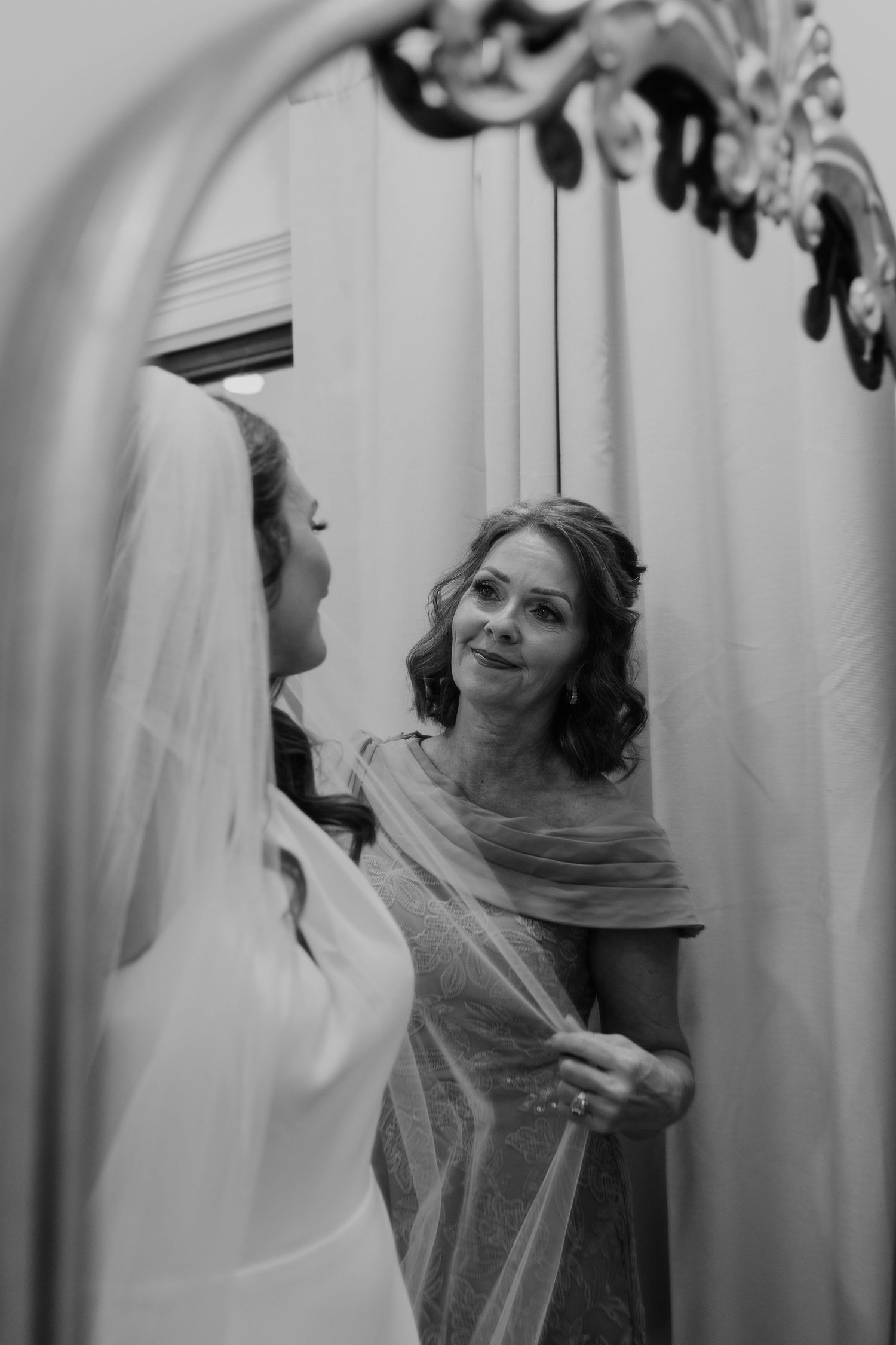 Black and white photo of a bride in veil with smiling woman adjusting her dress reflected in ornate mirror.