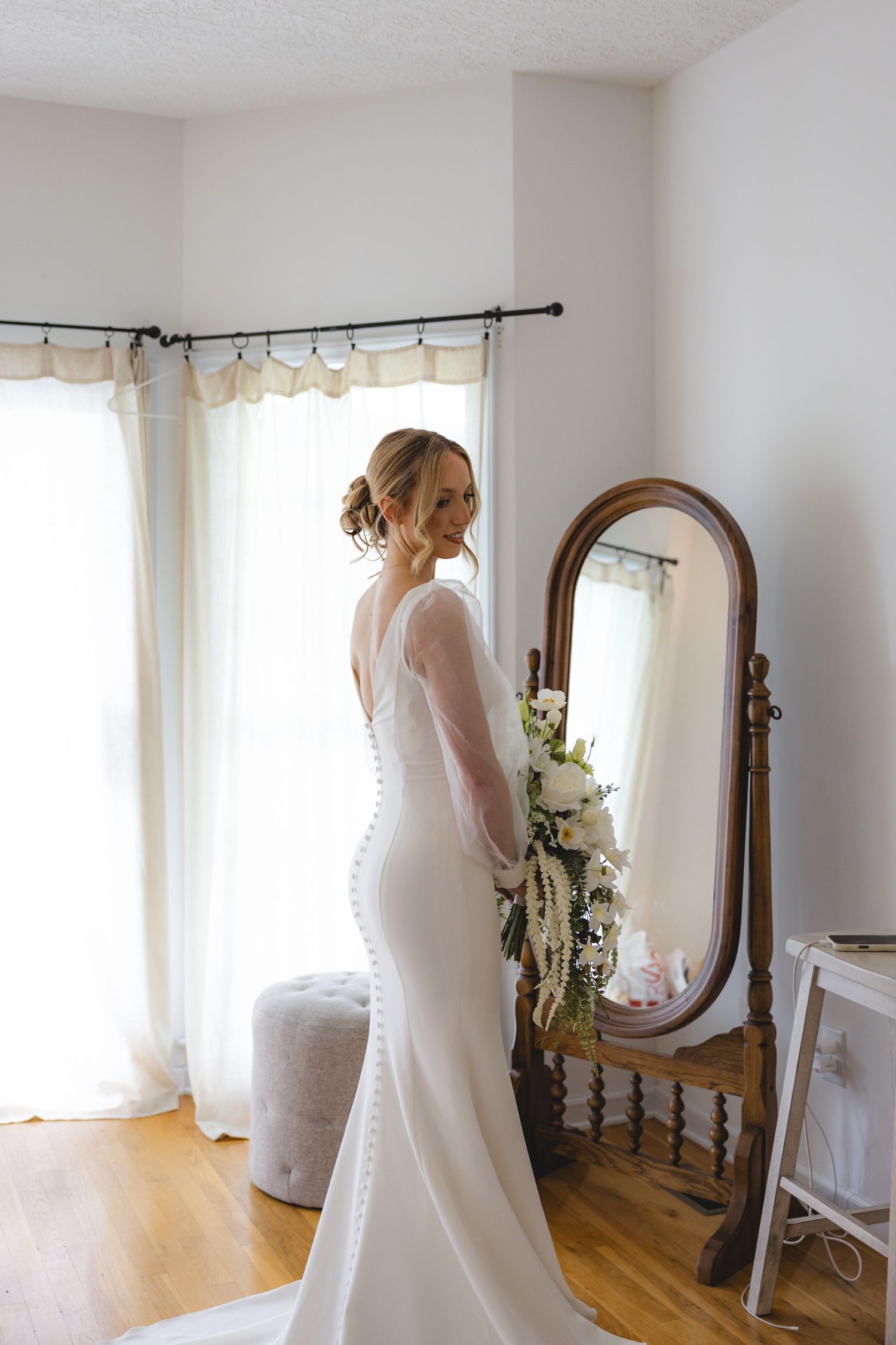 Bride in white mermaid gown holding bouquet, standing before ornate wooden floor mirror in bright room.