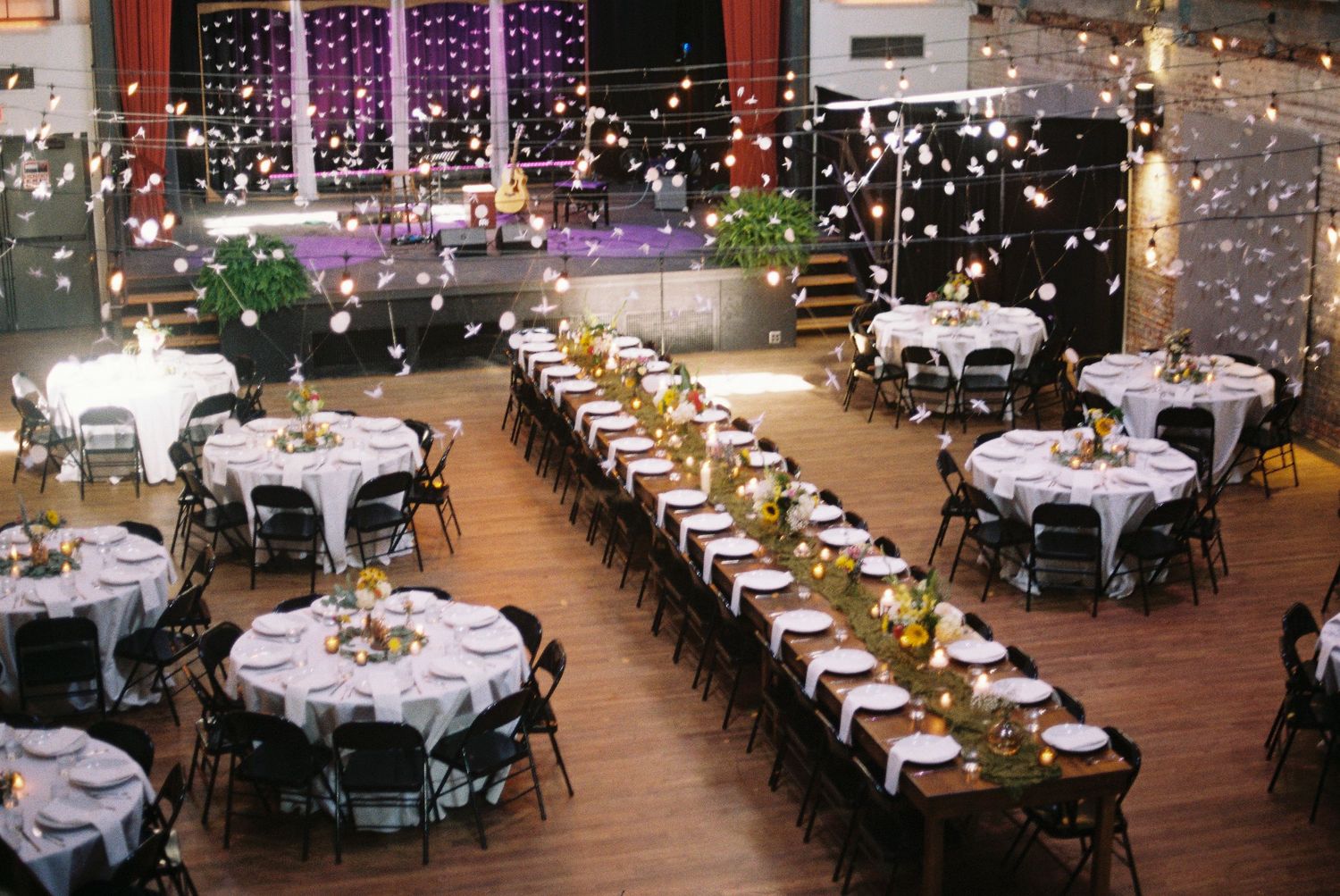 Elegant wedding reception venue with round and long tables, white linens, string lights, and bokeh lighting in a spacious hall.