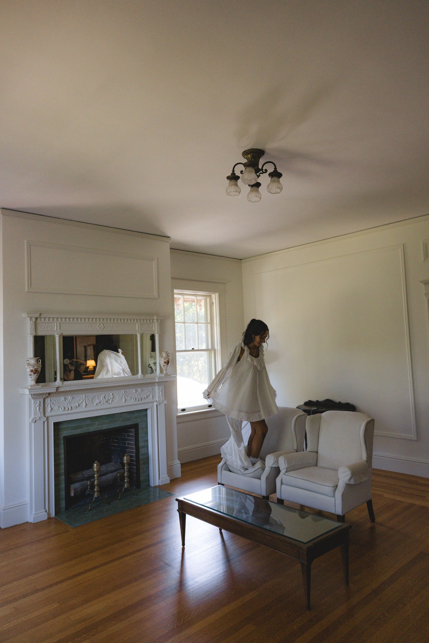 Woman in white dress standing on armchair in elegant living room with fireplace and hardwood floors.