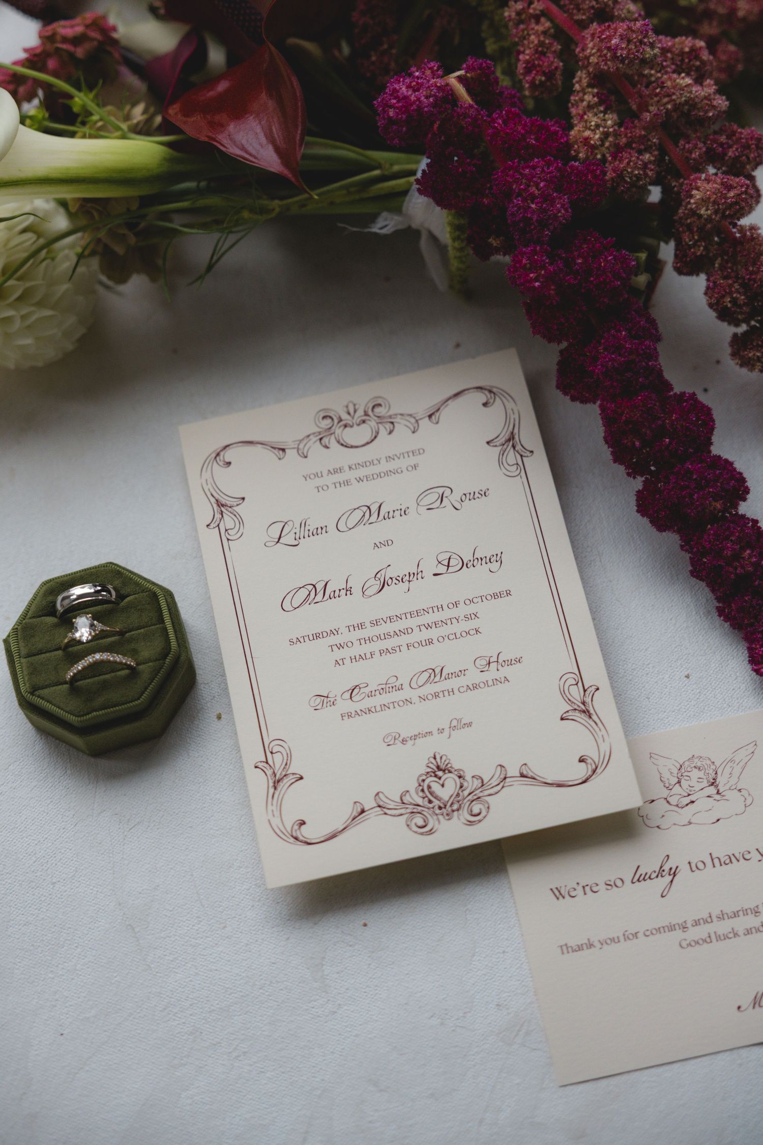 Cream wedding invitation with burgundy ornate border beside a green velvet ring box with wedding bands and deep magenta amaranth flowers.