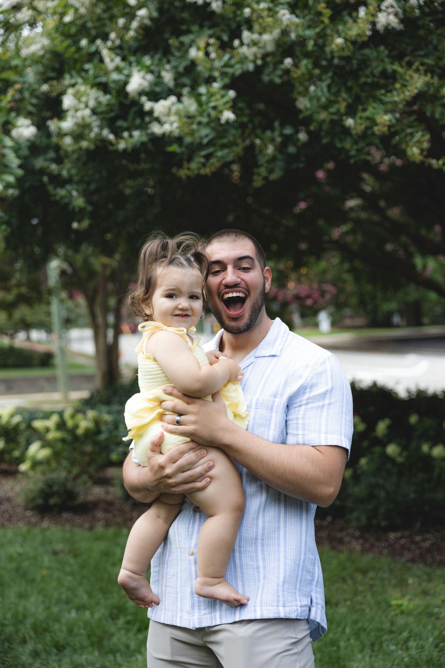 Laughing man holding a smiling baby girl in a yellow dress outdoors near blooming white flowers.