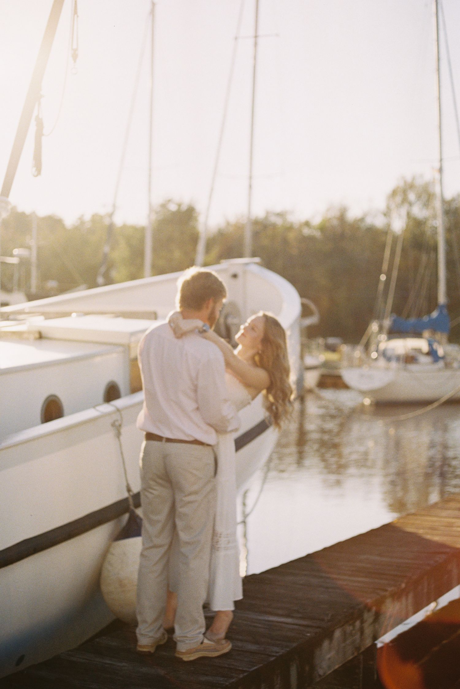 Couple embracing on a wooden dock at golden hour with sailboats in a marina behind them.
