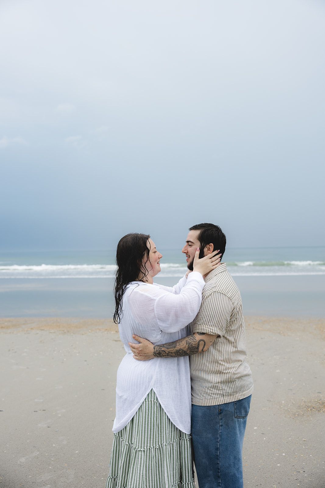 Couple embracing and smiling at each other on a sandy beach with ocean waves and cloudy sky behind them.