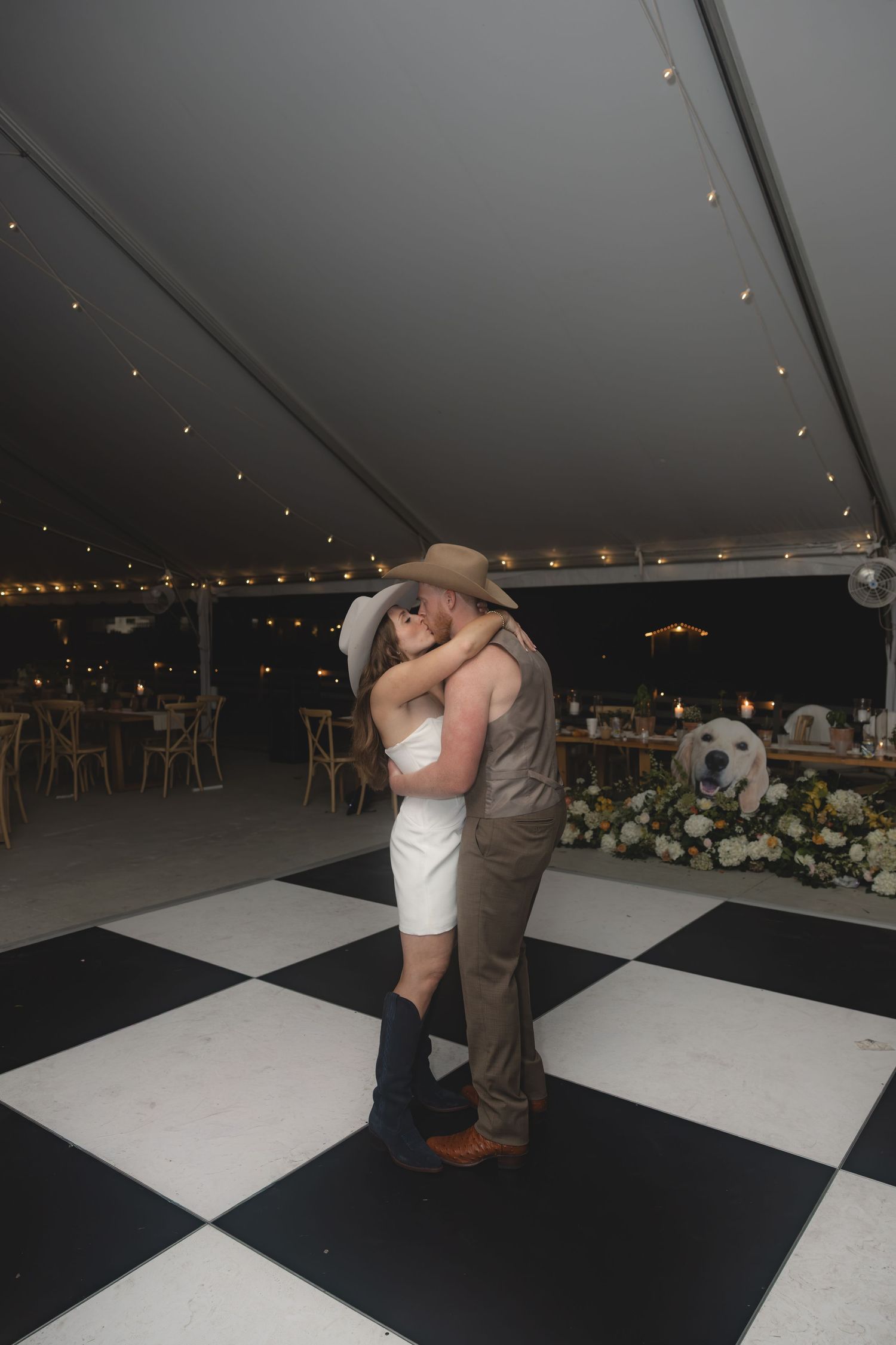 Bride and groom sharing a romantic kiss on a black and white checkered dance floor under a wedding tent.