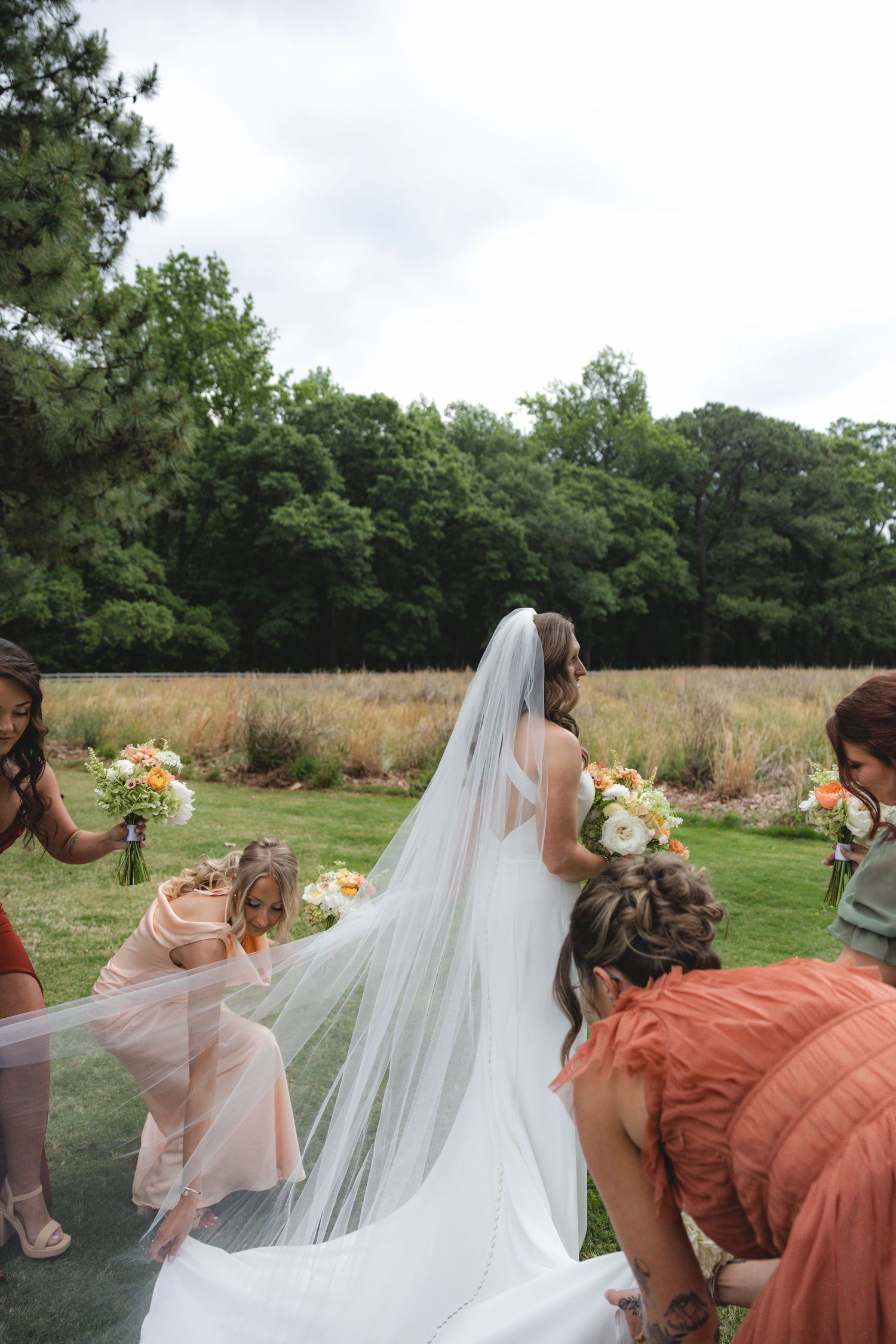 Bridesmaids adjust bride's long cathedral veil on green lawn before outdoor wedding ceremony.