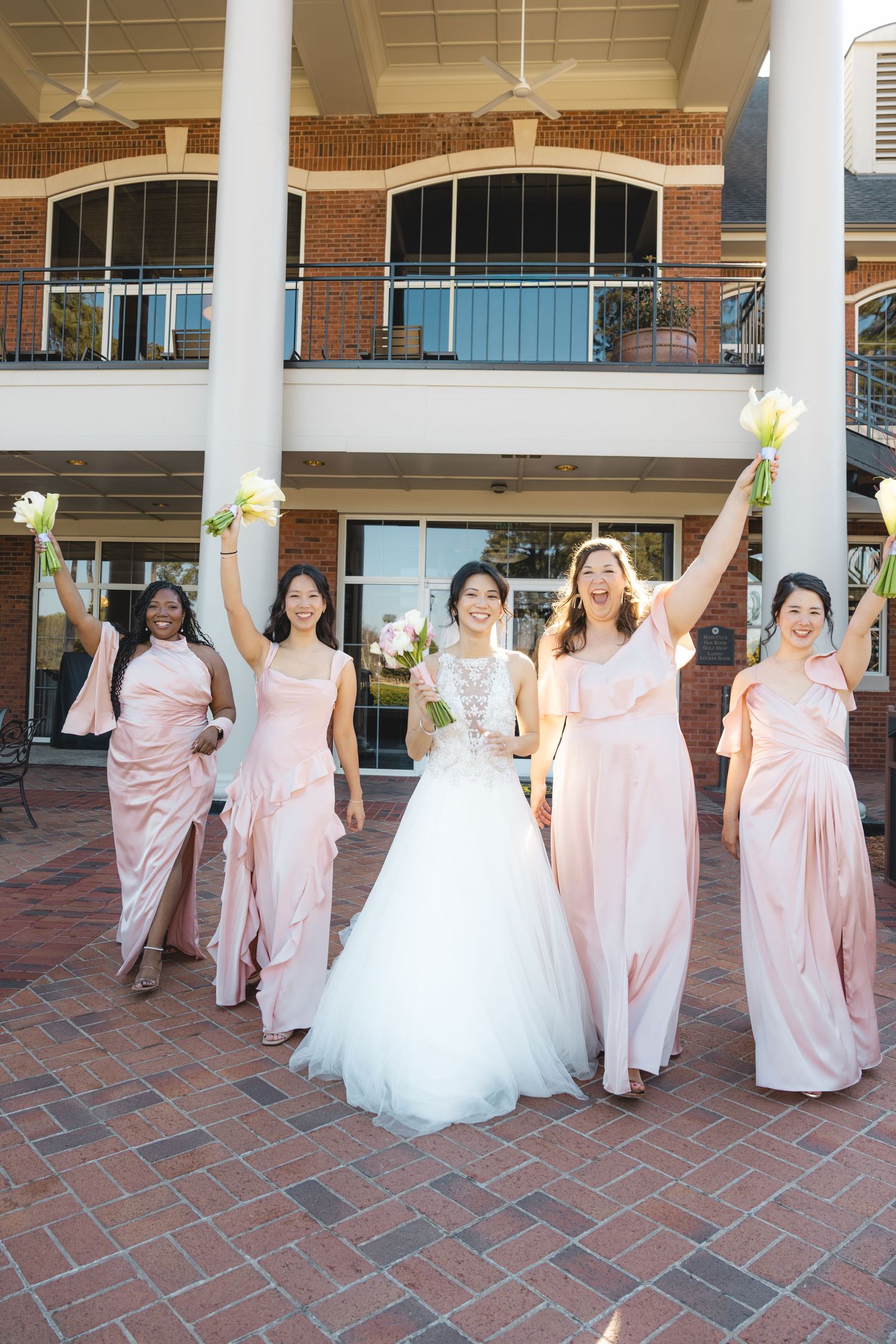 Bride in white ball gown celebrating with four bridesmaids in blush pink dresses raising bouquets.