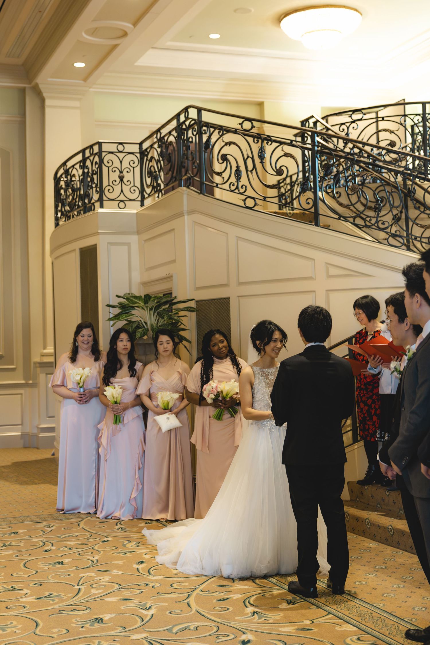 Bride and groom exchange vows before bridesmaids in dusty pink gowns near grand staircase.