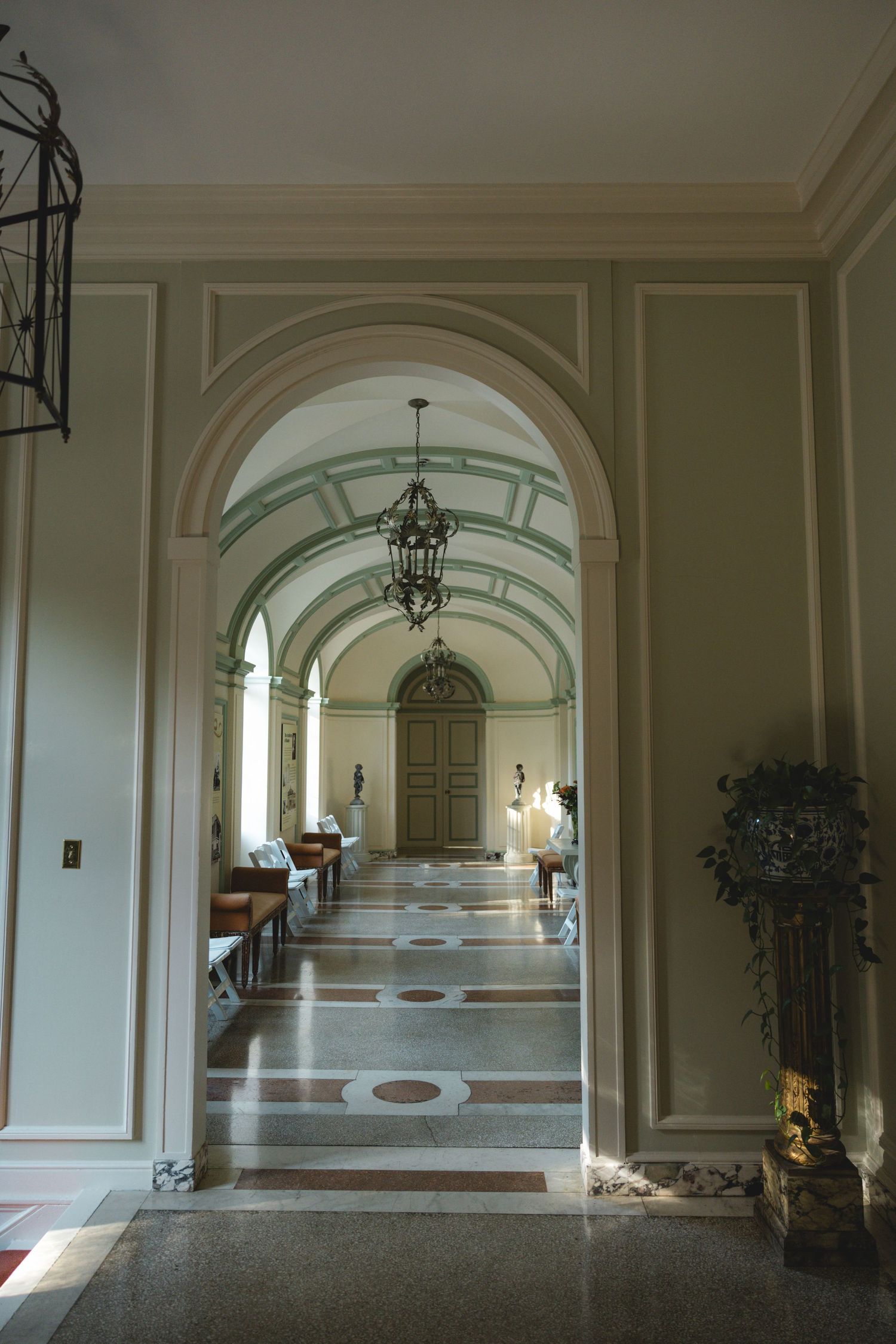 Elegant hallway with arched ceilings, ornate chandeliers, marble floors, and classical architectural details leading to a wooden door.