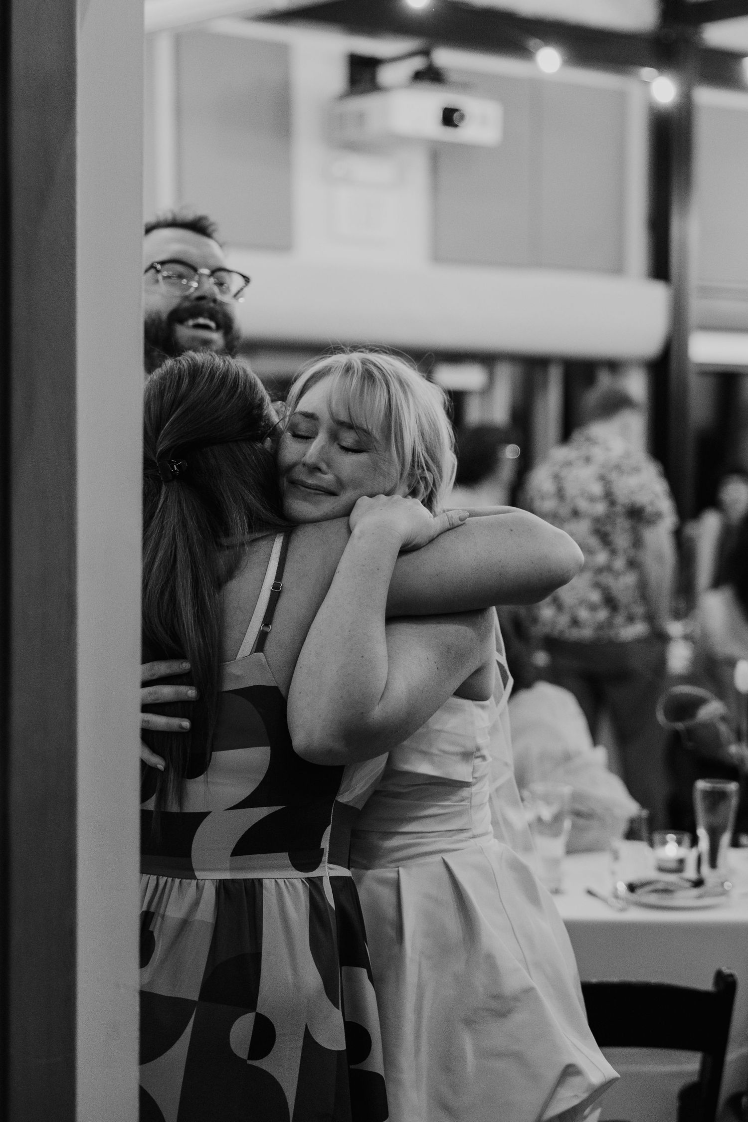 Black and white photo of two women sharing a warm embrace at a social gathering, with a smiling bearded man visible behind them.