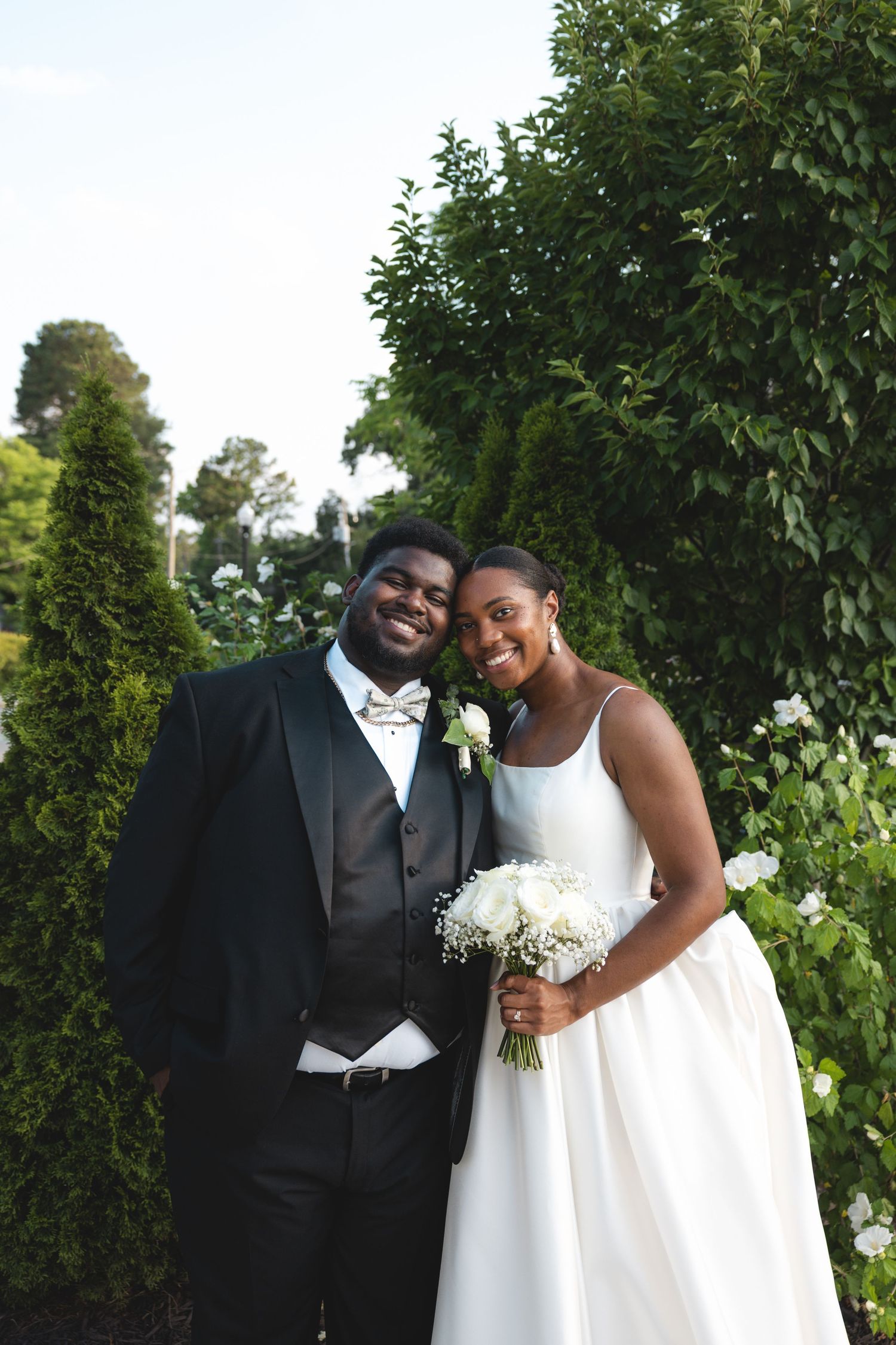 A joyful Black bride and groom pose outdoors in a lush garden, she in a white gown holding a bouquet, he in a black tuxedo.