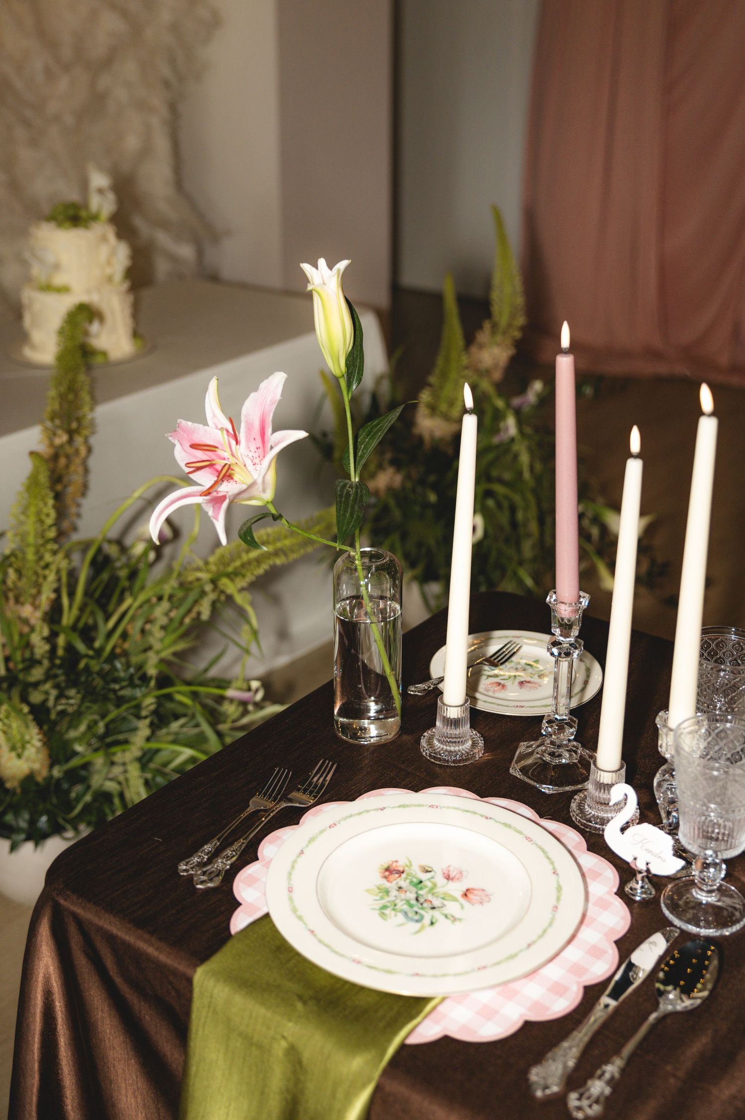Elegant wedding table setting with pink floral plate, taper candles, green runner, and pink lily centerpiece on brown satin tablecloth.
