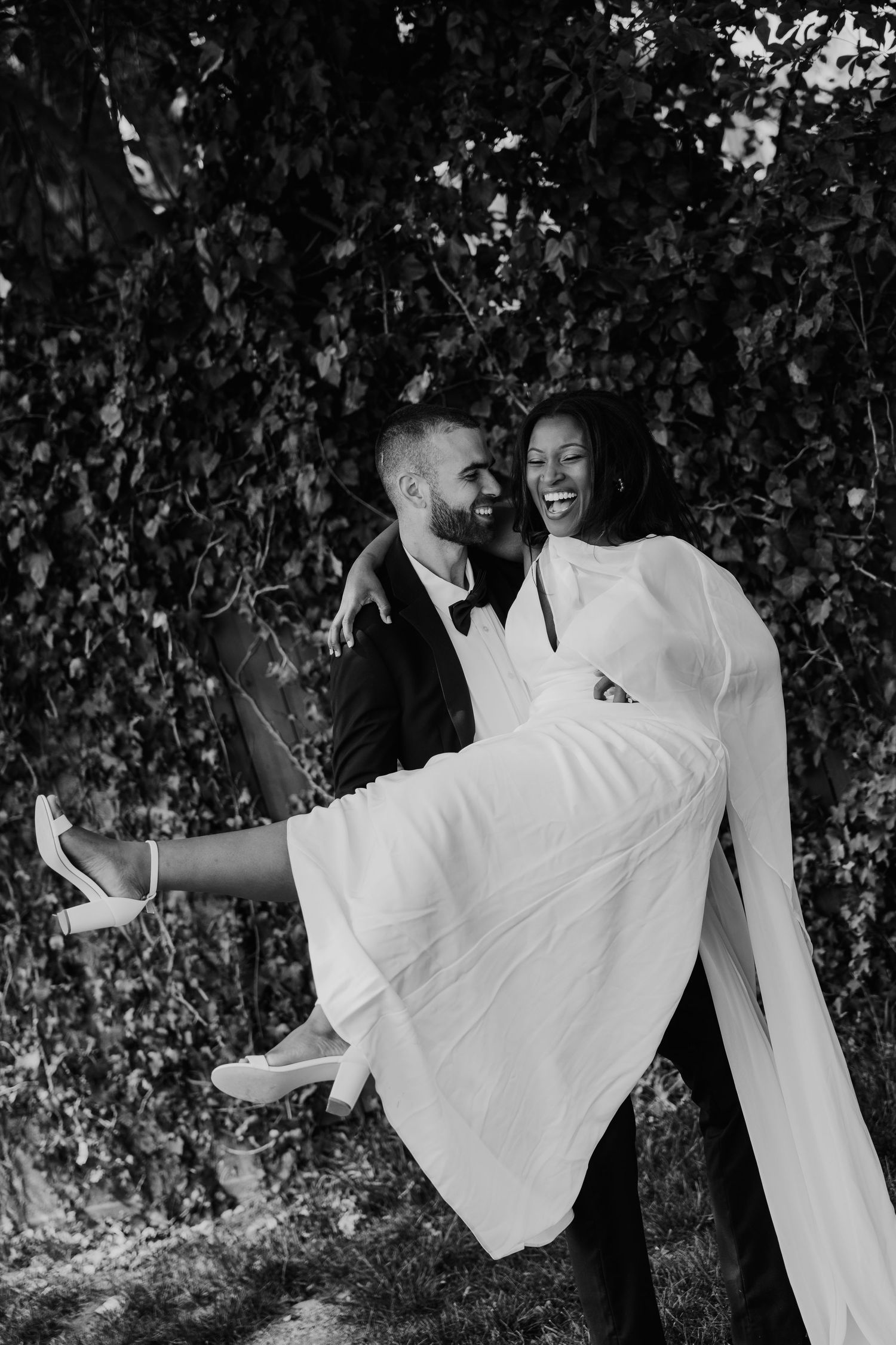 Black and white photo of a groom carrying his bride in a white gown, both smiling joyfully outdoors.