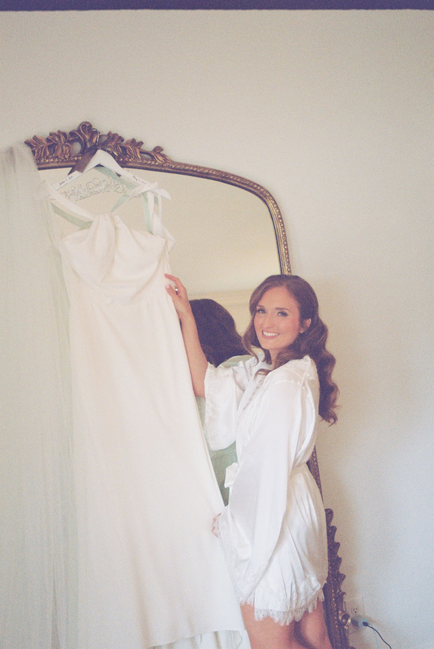Smiling bride in white robe holding her elegant wedding dress beside a gold ornate mirror.