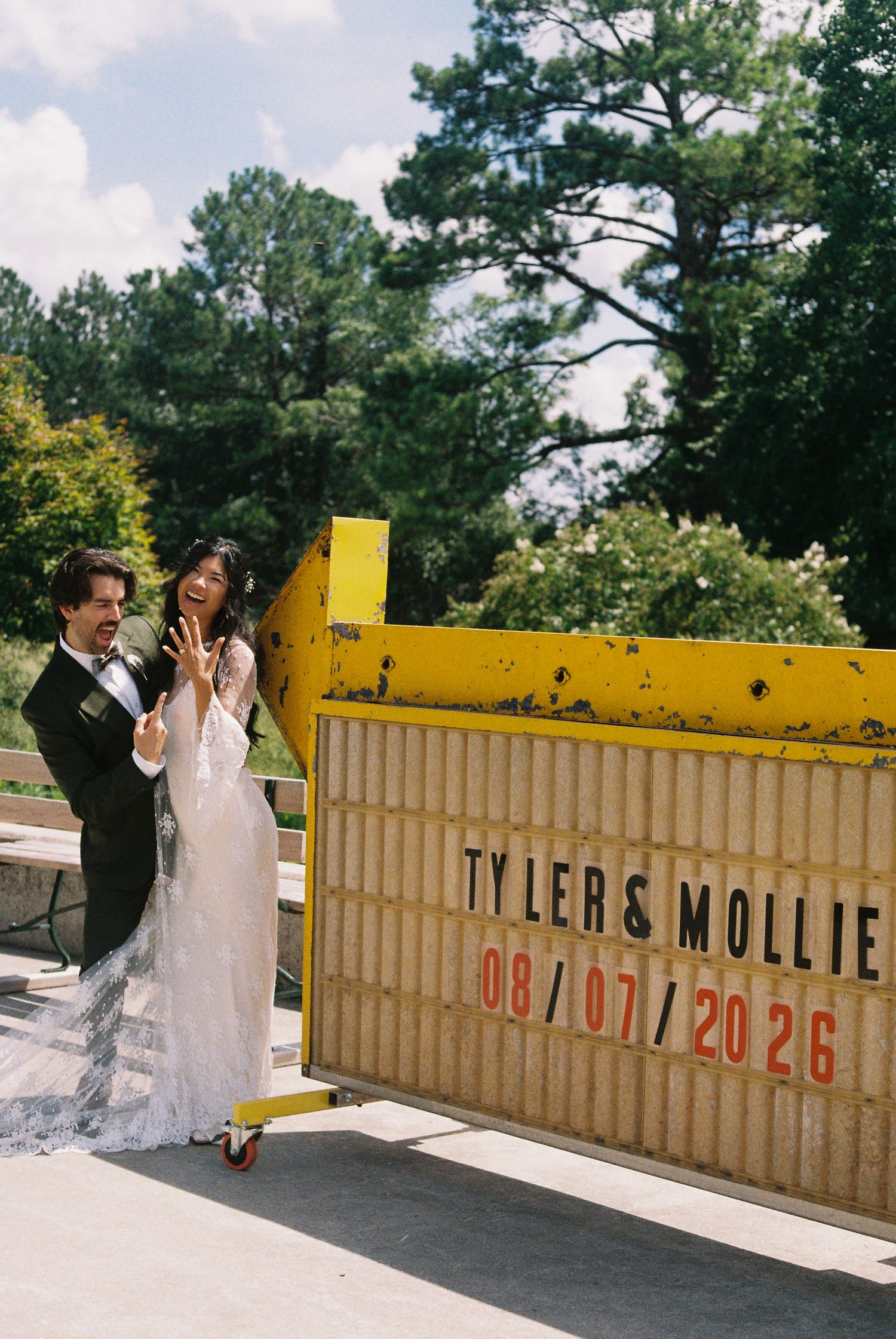 Bride and groom pose beside a yellow marquee sign reading Tyler & Mollie 08/07/2026 outdoors.