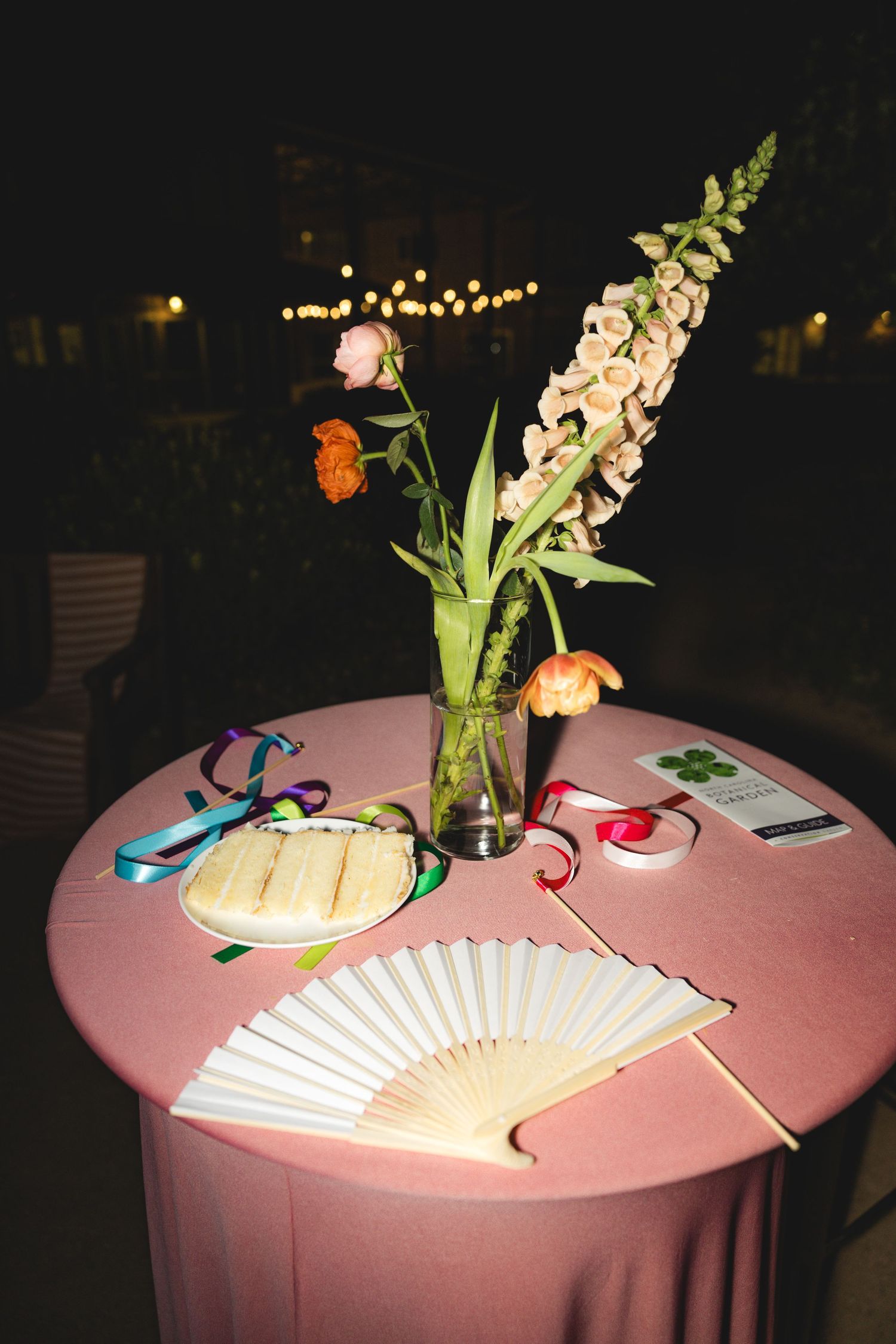 Elegant outdoor event table with pink cloth, floral centerpiece, white fan, ribbons, and party favors at night.