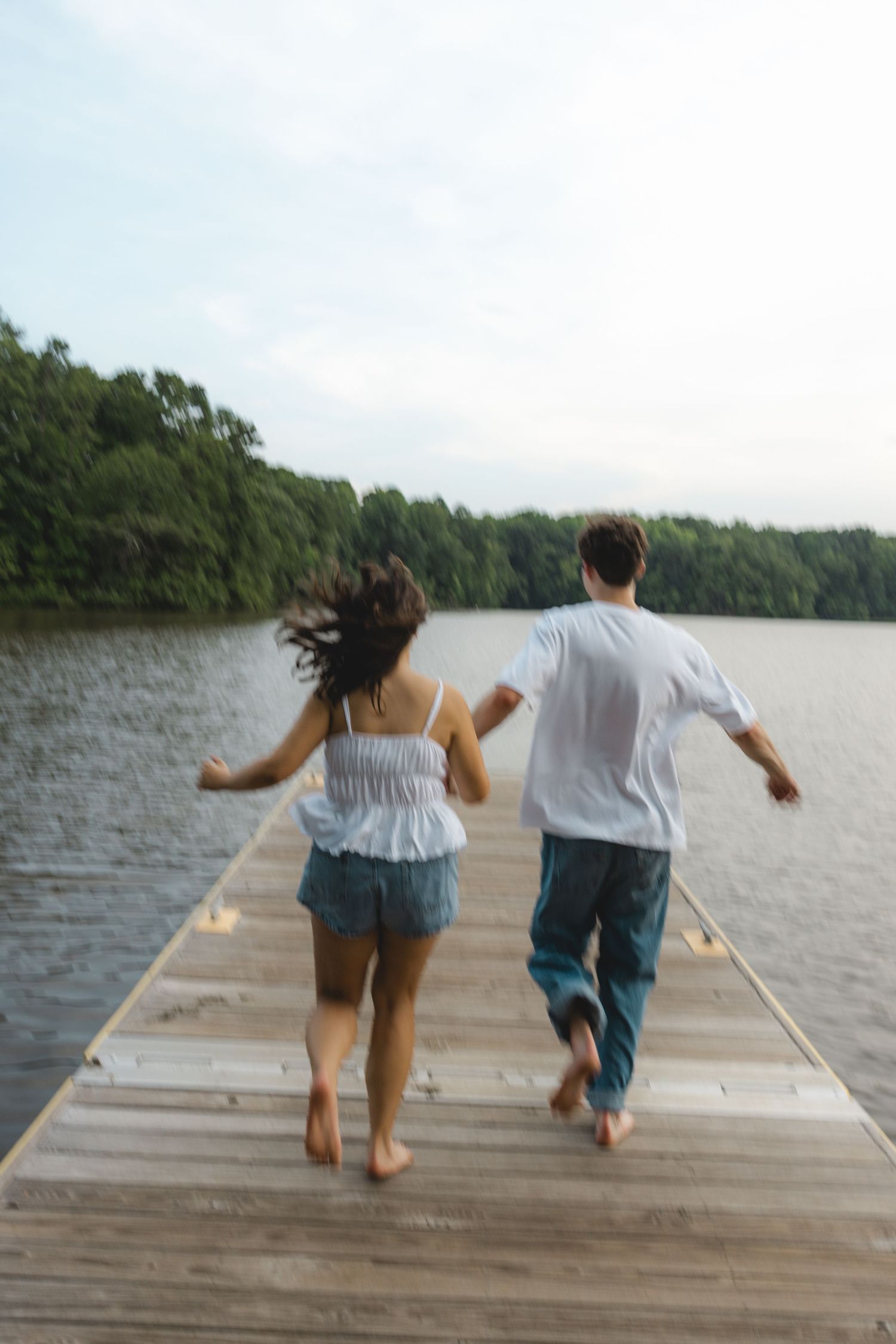 Couple running barefoot on wooden dock toward lake surrounded by lush green trees.
