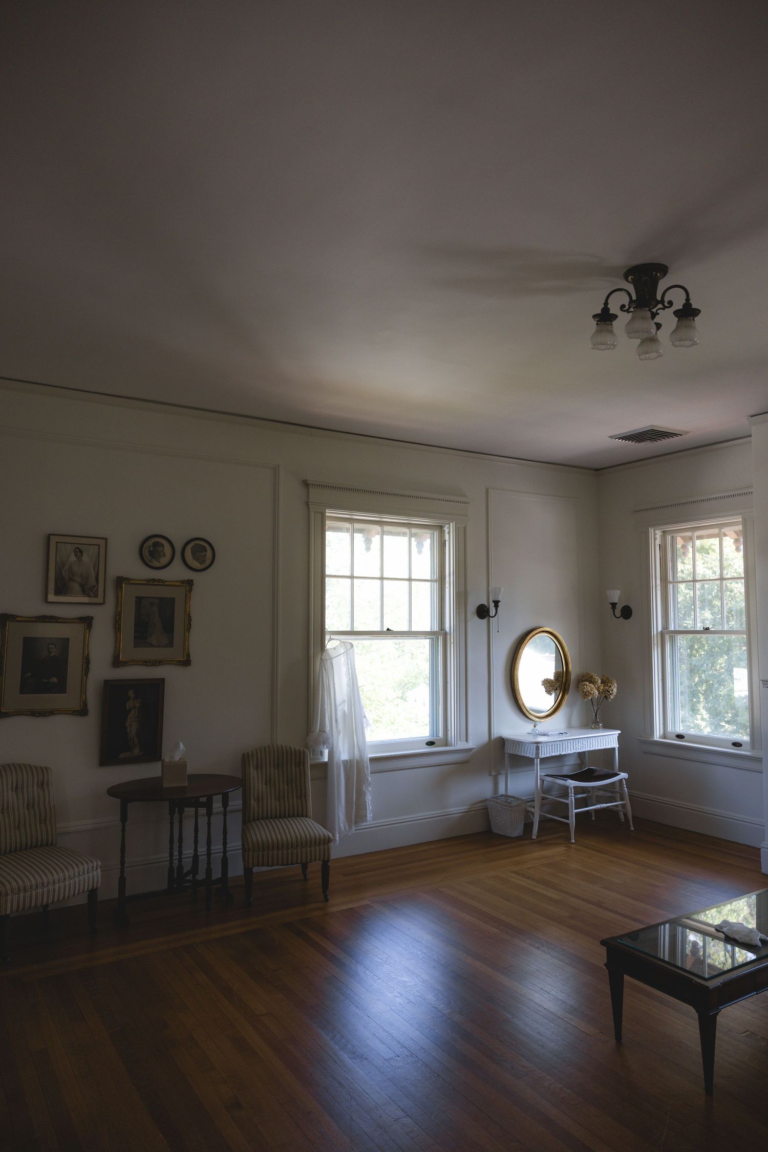 Vintage room with hardwood floors, white walls, oval mirror, vanity table, framed art, and natural light from two windows.