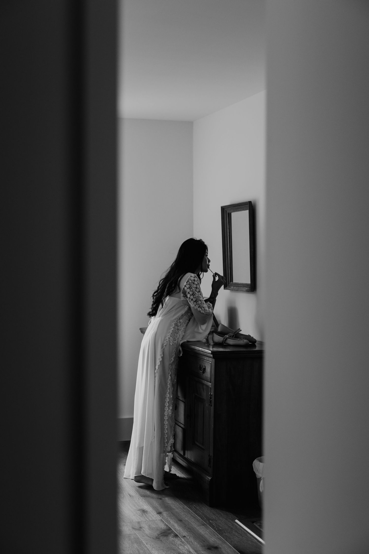 Black and white photo of a bride in a white gown standing at a dresser with mirror, getting ready on her wedding day.