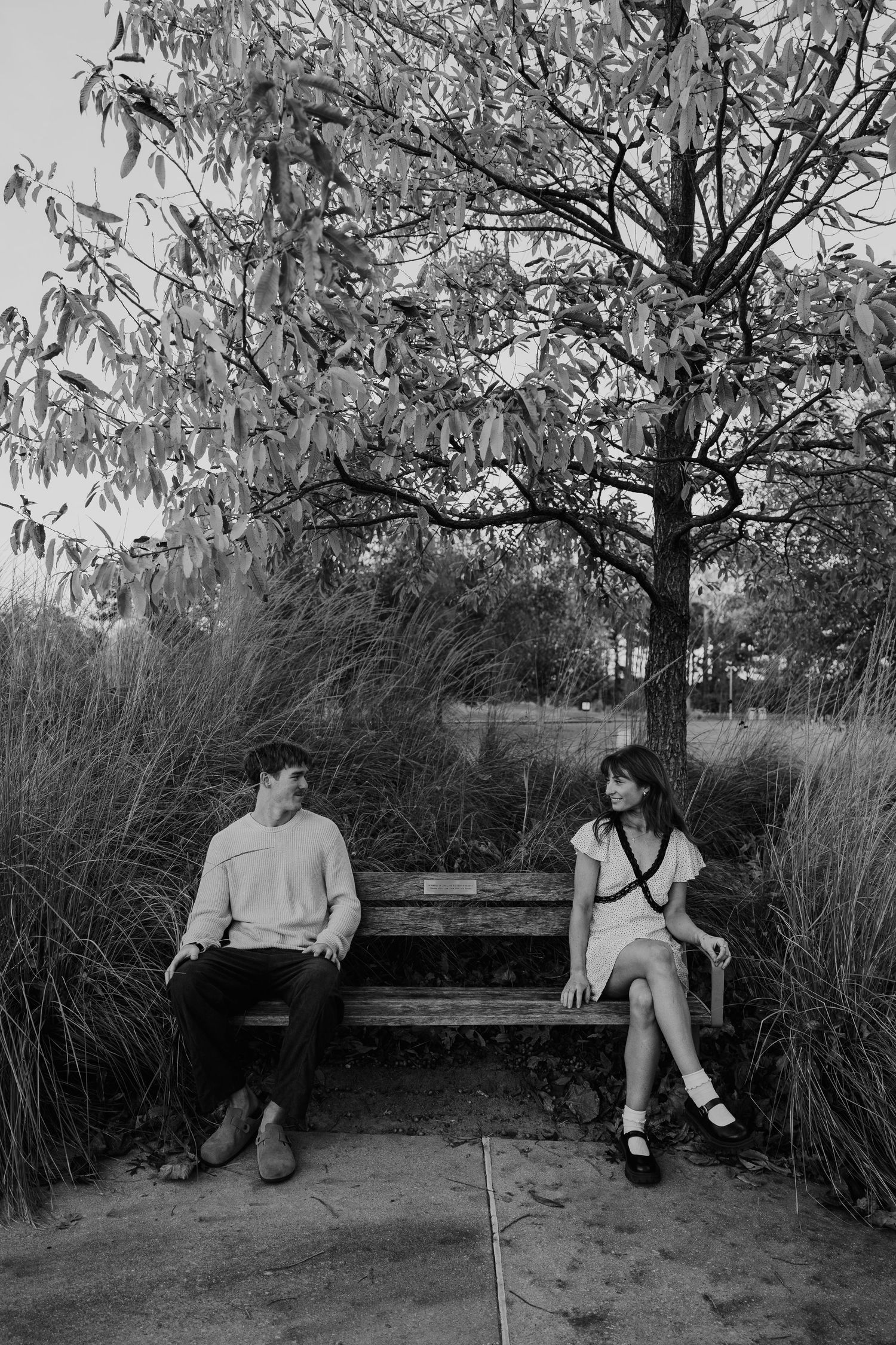 A couple sits apart on a wooden bench surrounded by trees in black and white.