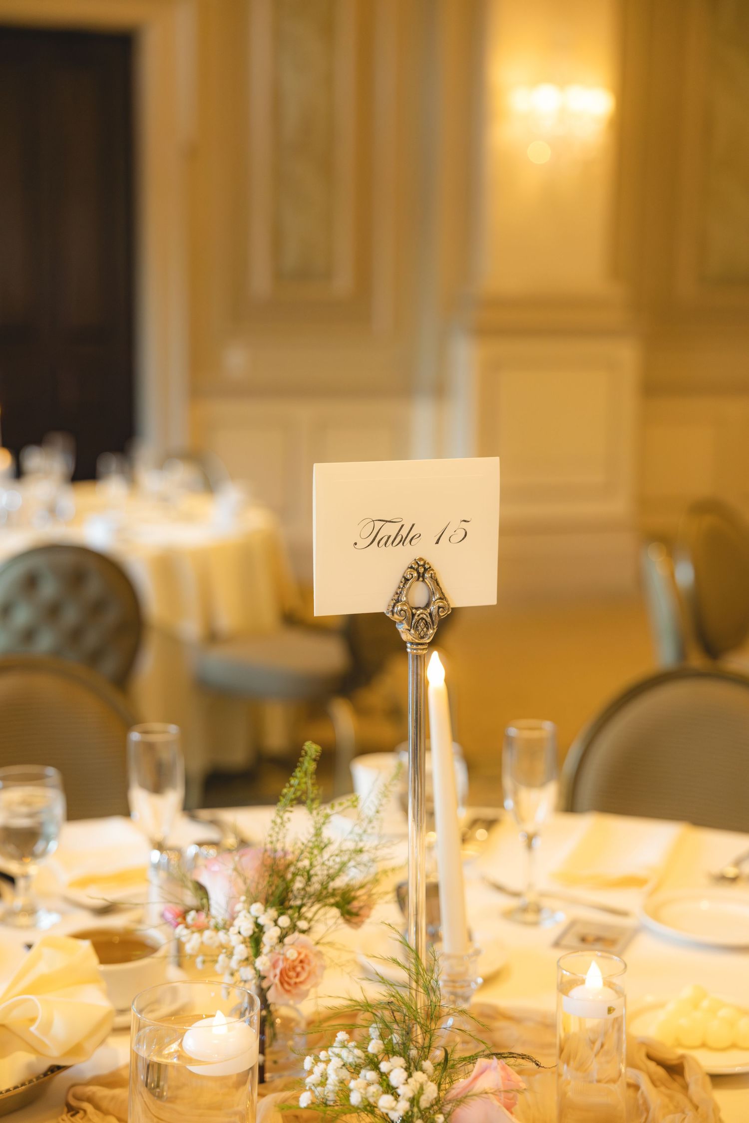 Elegant wedding reception table number 15 card in ornate holder with pink roses and candles in grand ballroom.